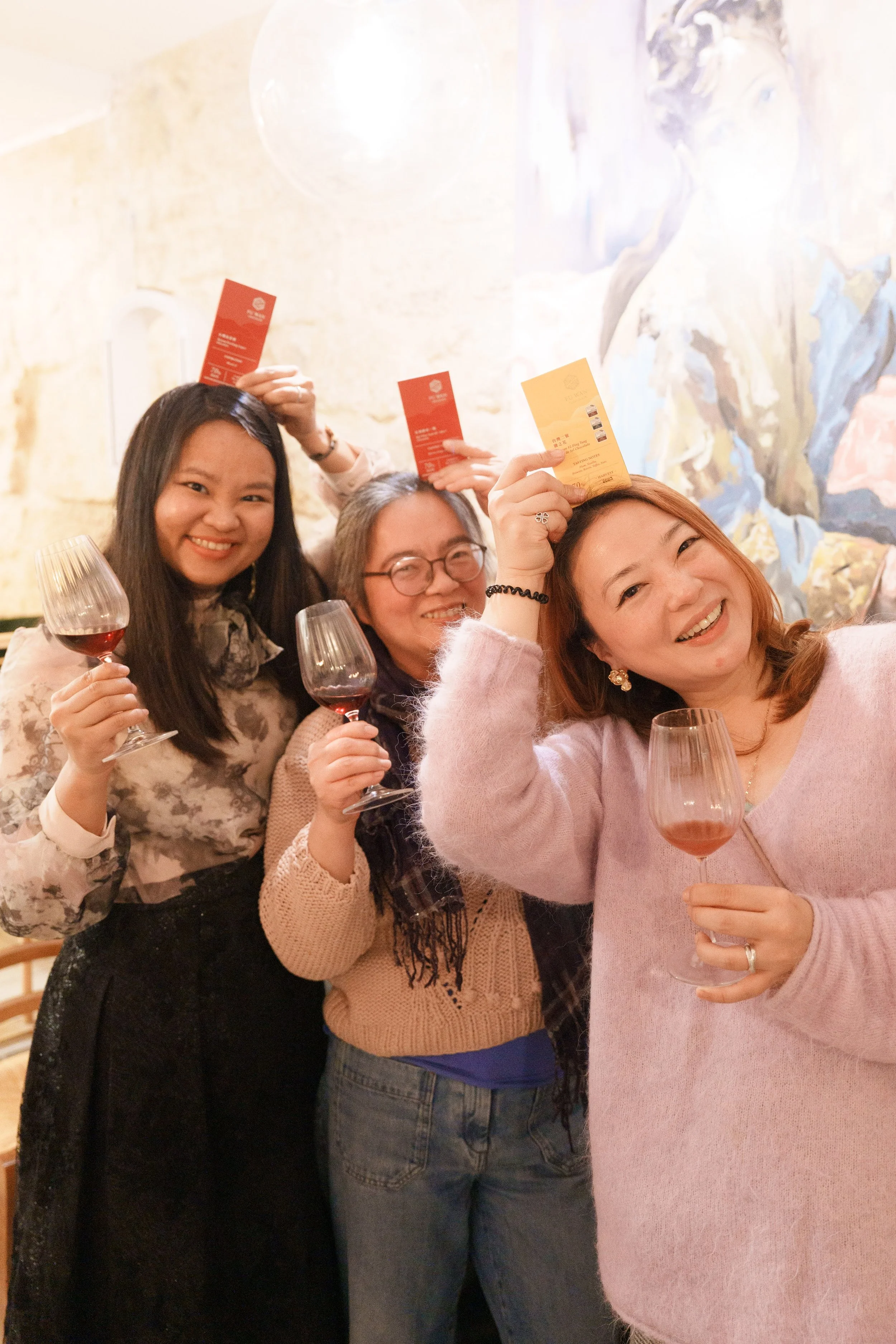 Trois femmes souriantes assises autour d'une table, tenant des verres de vin et des tablettes de chocolats, lors d'un événement dans un restaurant avec un fond mural artistique.