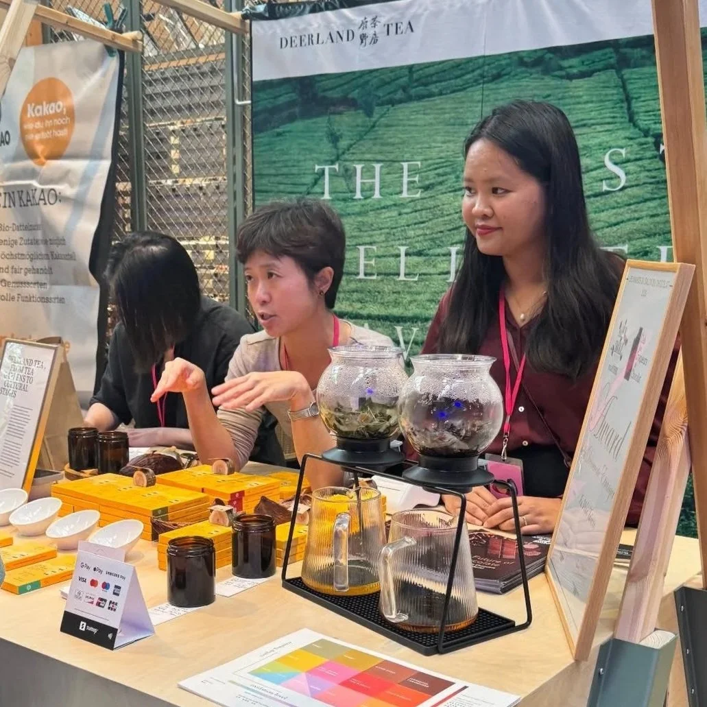 trois femmes dans un stand de marché culinaire avec du chocolat et du thé specialité, ainsi qu'un grand panneau de fond vert d'un jardin du thé taïwanais.