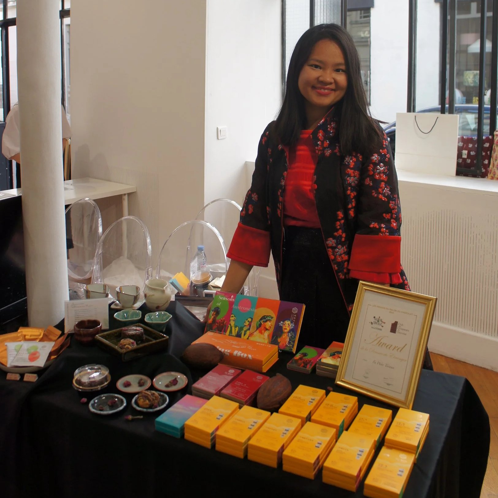 Femme souriante debout derrière un étalage de produits dans une boutique ou un marché intérieur, avec des articles comme des tasses, des chocolat tree-to-bar et des coffrets, dans un espace bien éclairé avec des grandes fenêtres.