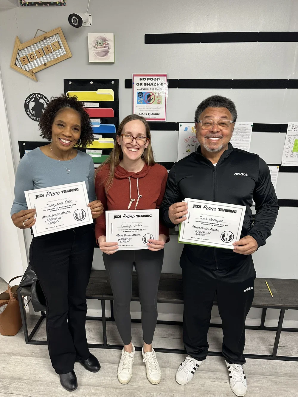 Three people standing indoors, each holding a certificate of completion for a piano training class, smiling at the camera after music lessons.