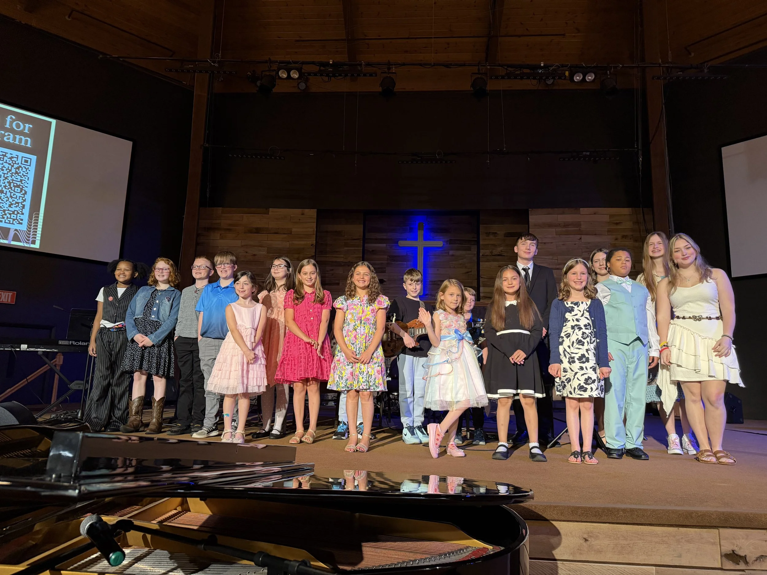 A group of children and teenagers standing on stage inside a church, with a cross on the wall behind them. There is a blue glow around the cross and a young girl playing guitar is visible among them. Two large screens display QR codes and text on either side of the stage.