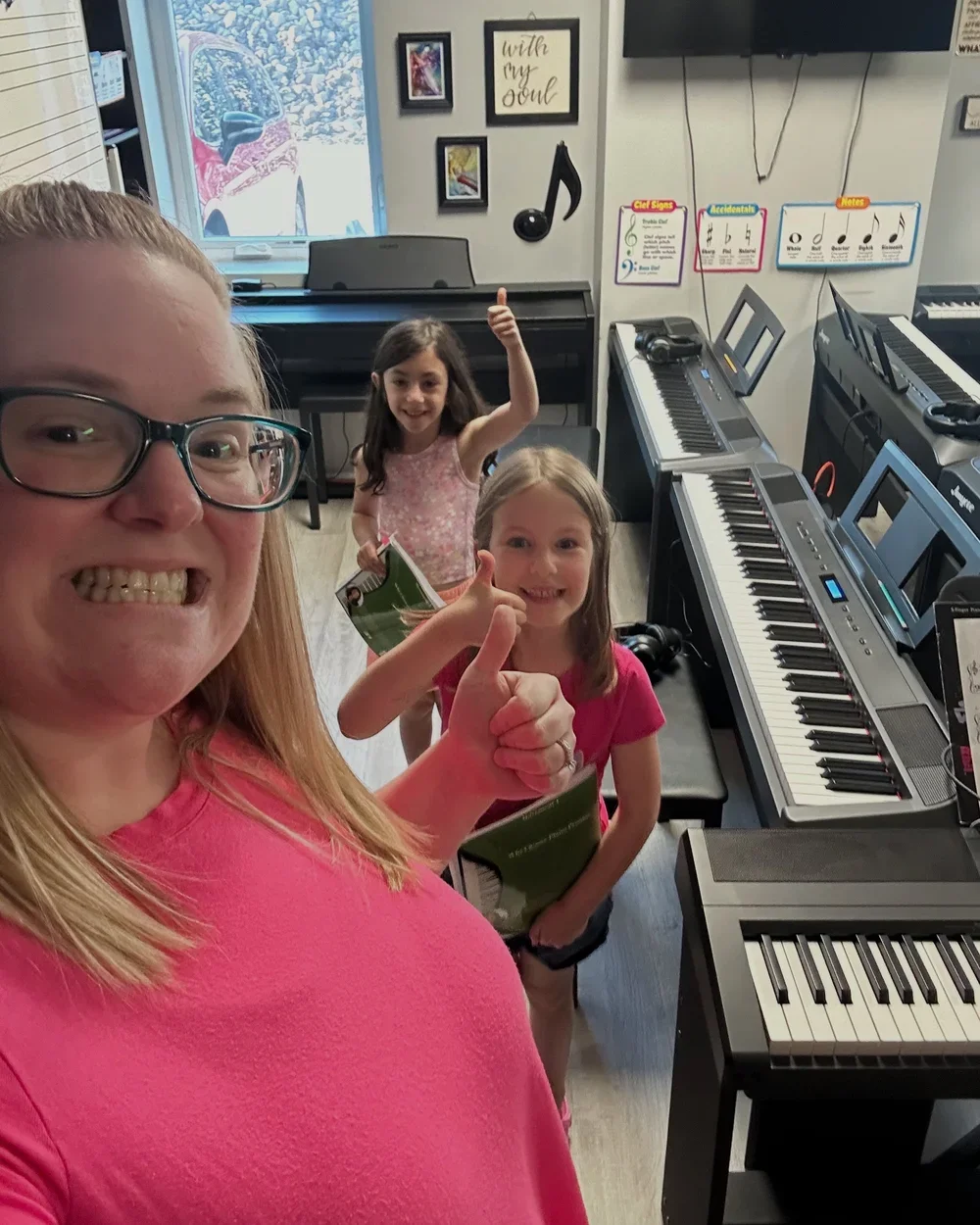 A woman and two young girls smiling in a music store surrounded by electronic keyboards. The woman is giving a thumbs up and smiling broadly. One girl in a pink shirt is giving a thumbs up, and the other girl in a pink floral top is smiling and givin