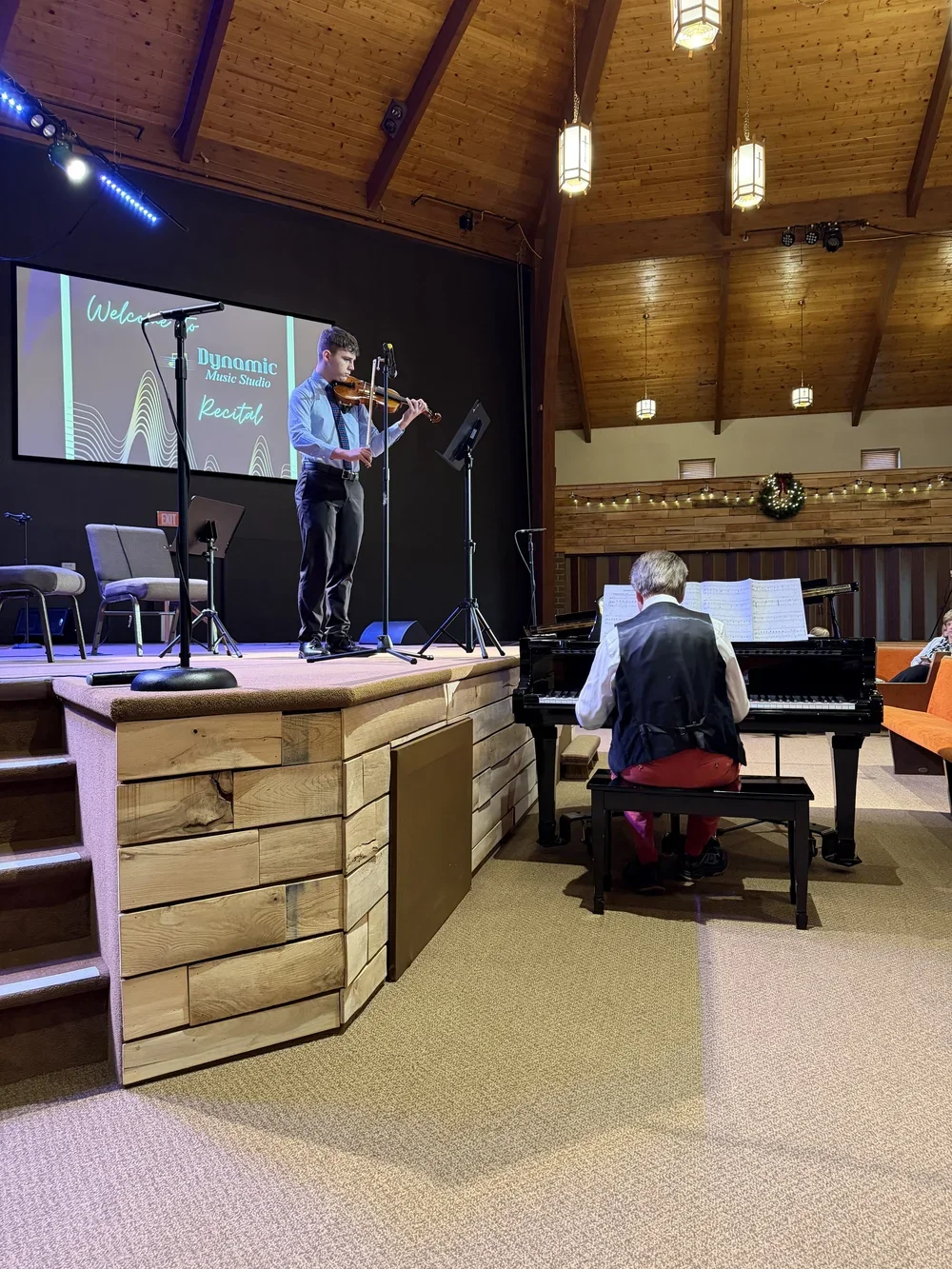 A young man playing the violin on stage with a pianist accompanying him. The stage features a large screen with the words "Dynamic Music Studio recital" at the venue for a Christmas recital.