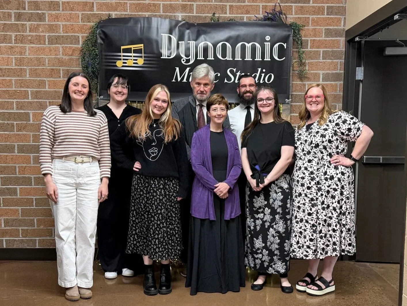 Group of nine people posing in front of a sign of a music lesson studio that reads 'Dynamic Music Studio' with musical notes on a brick wall background.
