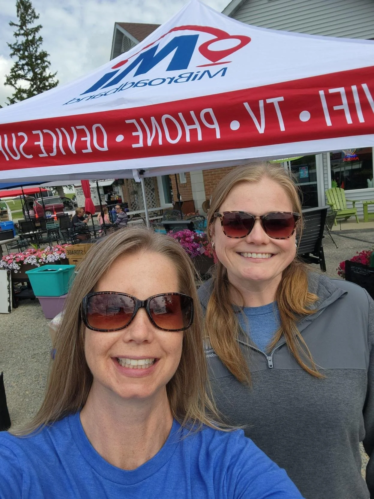 Two women smiling and taking a selfie outdoors beneath a white canopy with red and blue text and logo. One woman has long blonde hair and sunglasses, wearing a blue shirt; the other has long light brown hair and sunglasses, wearing a gray jacket. There are people, flowers, and outdoor seating in the background.