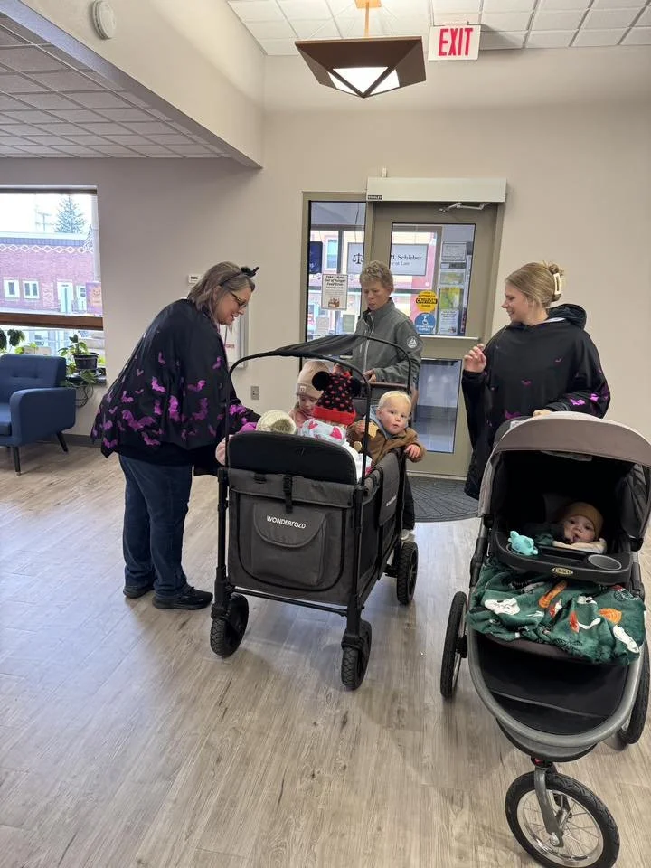 Two women and two children in strollers inside a building, near an exit door with glass panes. One woman is talking to the children in a stroller, while the other woman stands beside a baby in a stroller. The room has windows, a blue chair, and a wooden floor.
