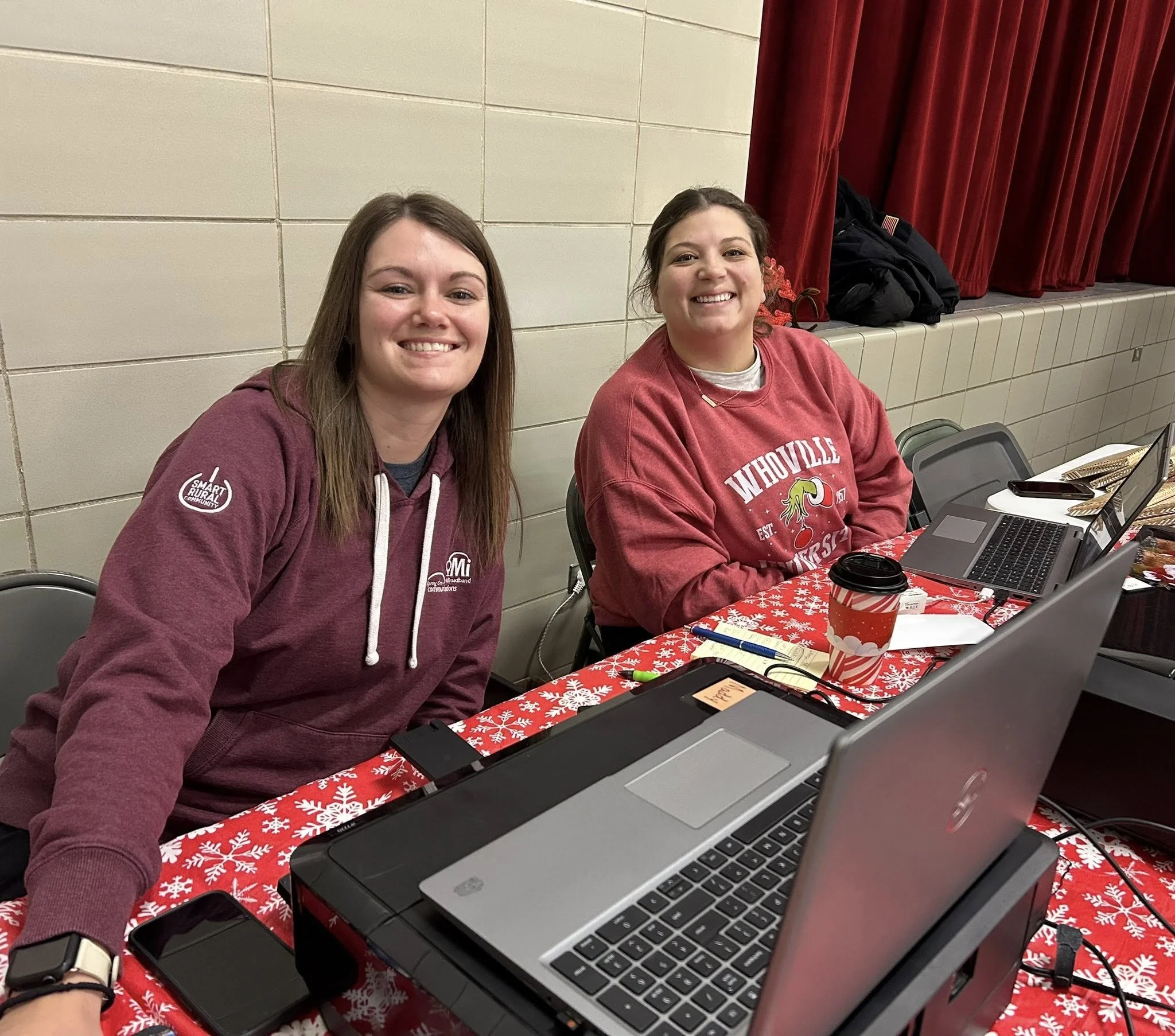 Two women sitting at a table with a red holiday-themed tablecloth, smiling at the camera. One woman is wearing a maroon hoodie and the other a red sweatshirt. There are laptops and a coffee cup on the table, and a black backpack is on a ledge behind them. Red curtains are in the background.