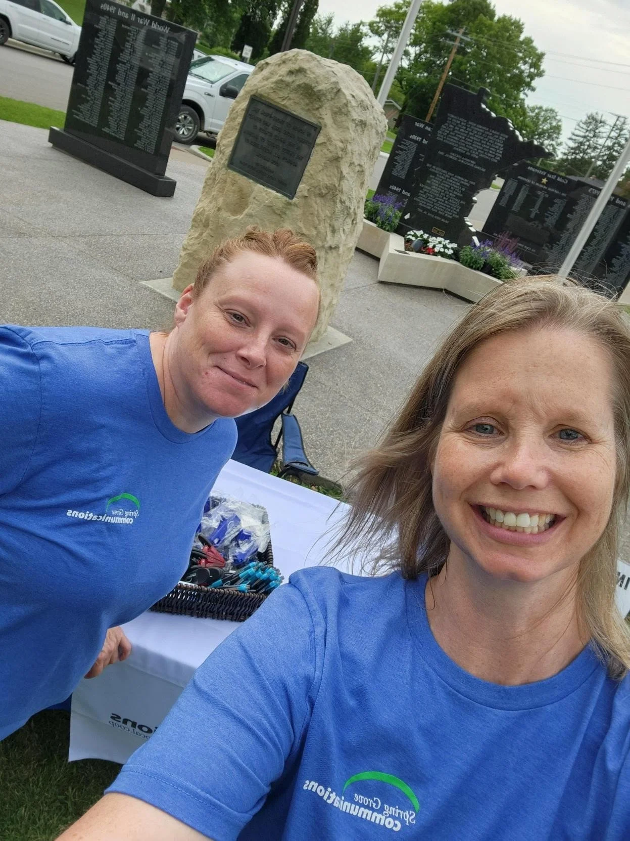 Two women wearing blue t-shirts taking a selfie outdoors near a table with various supplies, with memorial plaques and tombstones in the background.