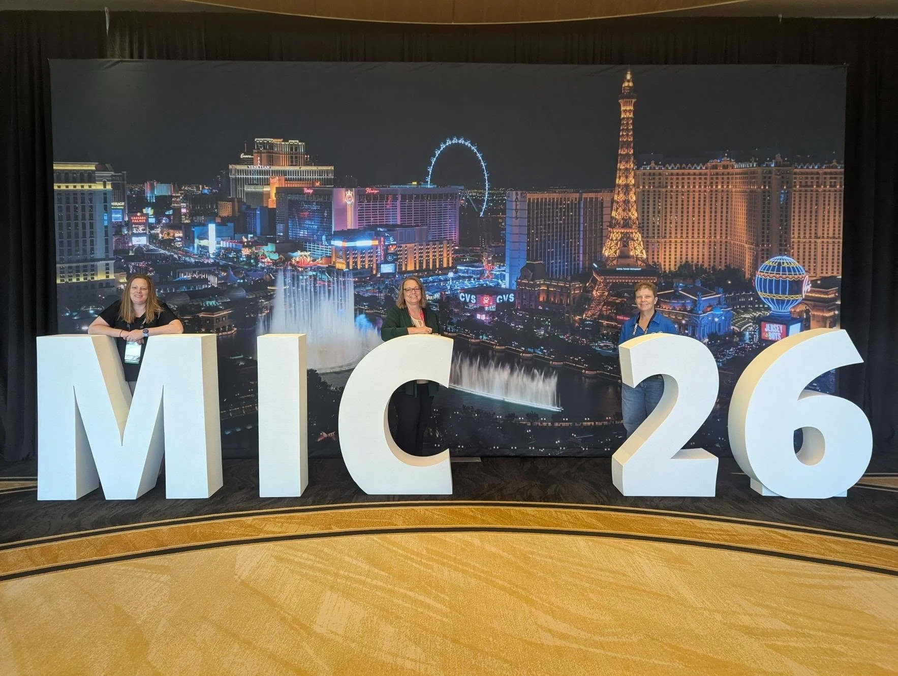 Three women standing behind large white 3D letters and numbers spelling out 'M C 26' in front of a Las Vegas backdrop with city lights and iconic landmarks, including the Eiffel Tower replica and a Ferris wheel, at night.