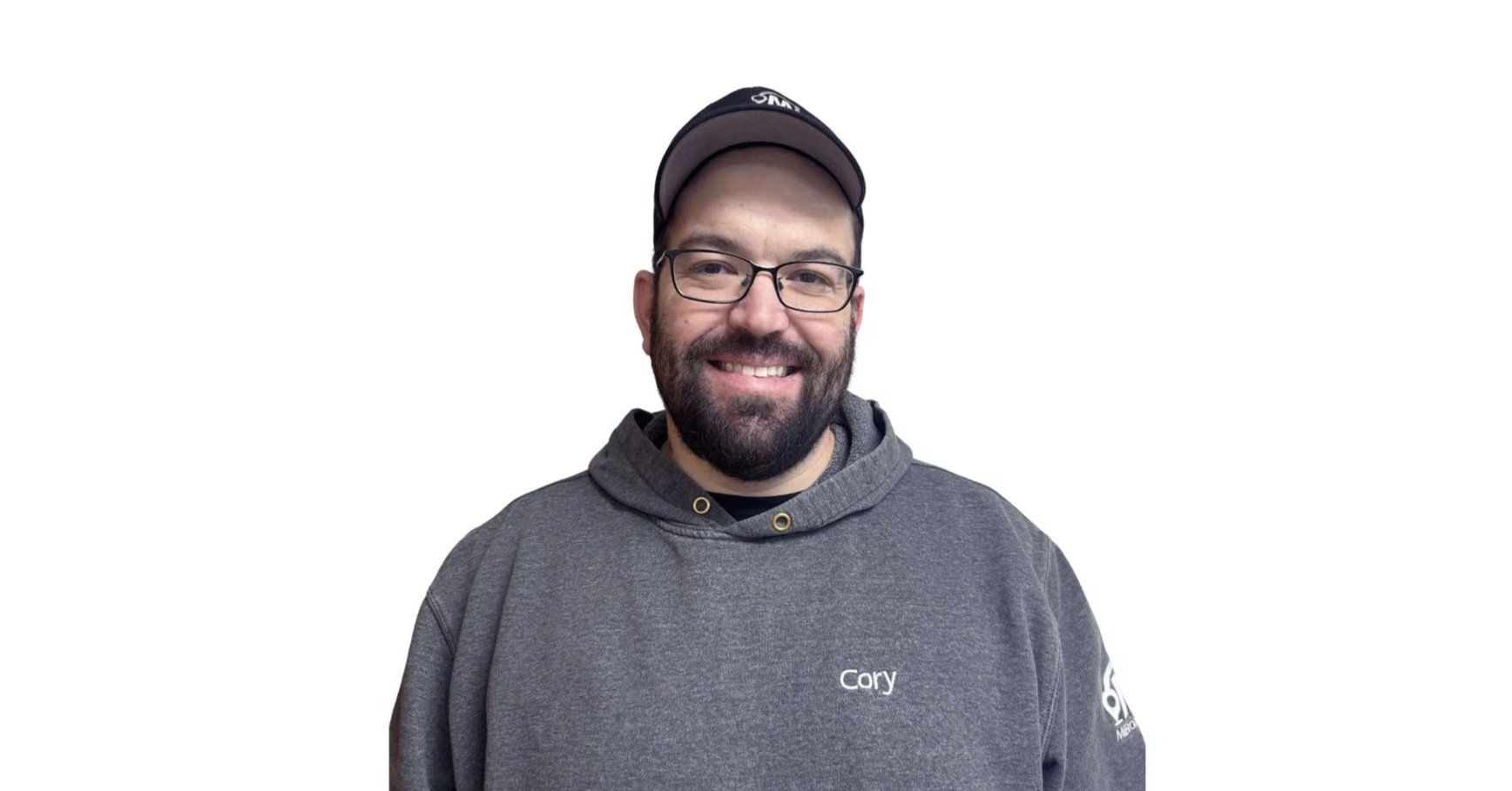 A man with glasses and a beard smiling, wearing a gray hoodie with "Cory" embroidered on it and a black cap, against a plain white background.