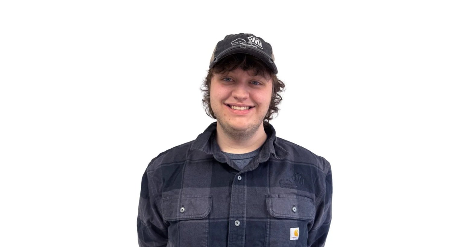 A young man with curly brown hair wearing a black cap and black Carhartt jacket, smiling against a white background.
