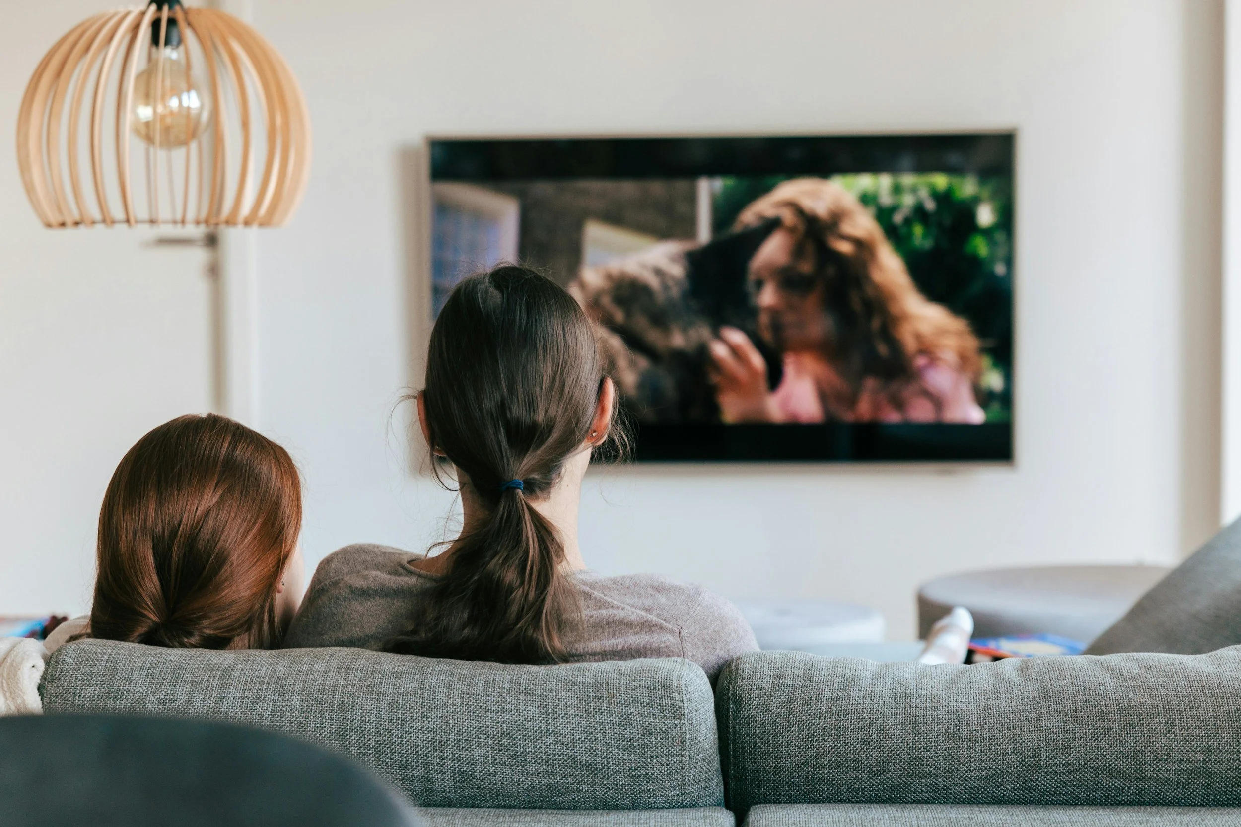 Three women sitting on a gray couch watching a television in a living room. The television shows a woman with long hair holding a dog. There is a wooden pendant light hanging from the ceiling to the left of the image.
