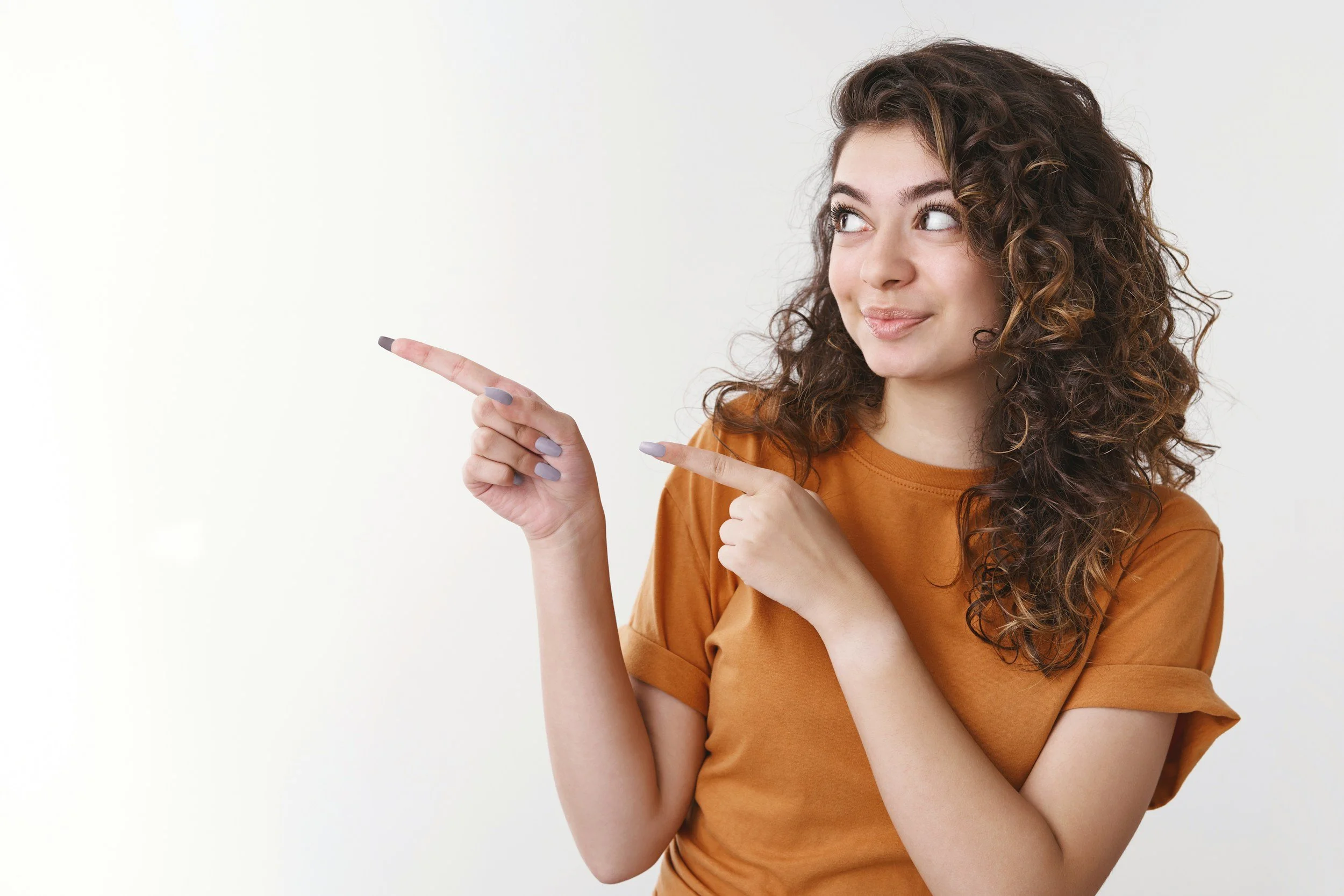 Young woman with curly brown hair wearing an orange shirt, pointing to her right with both index fingers, smiling softly, against a plain white background.