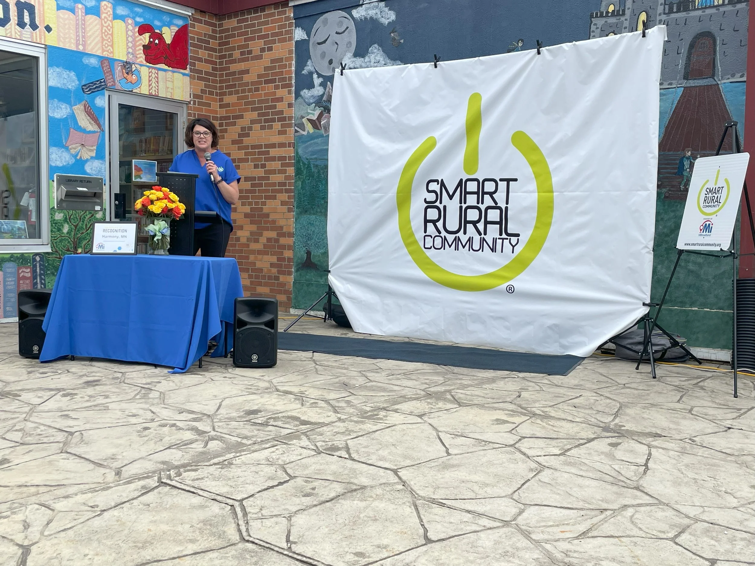 A woman with glasses wearing a blue shirt is standing on a stage, speaking into a microphone, in front of a large white banner with the words "Smart Rural Community" and a logo. There is a table with a flower arrangement, a sign, and some speakers. The background features a colorful mural on a brick building wall.