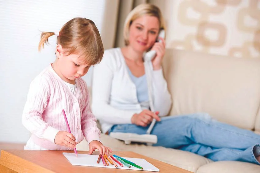 A young girl coloring at a table while a woman, possibly her mother, is sitting on a couch in the background talking on the phone.