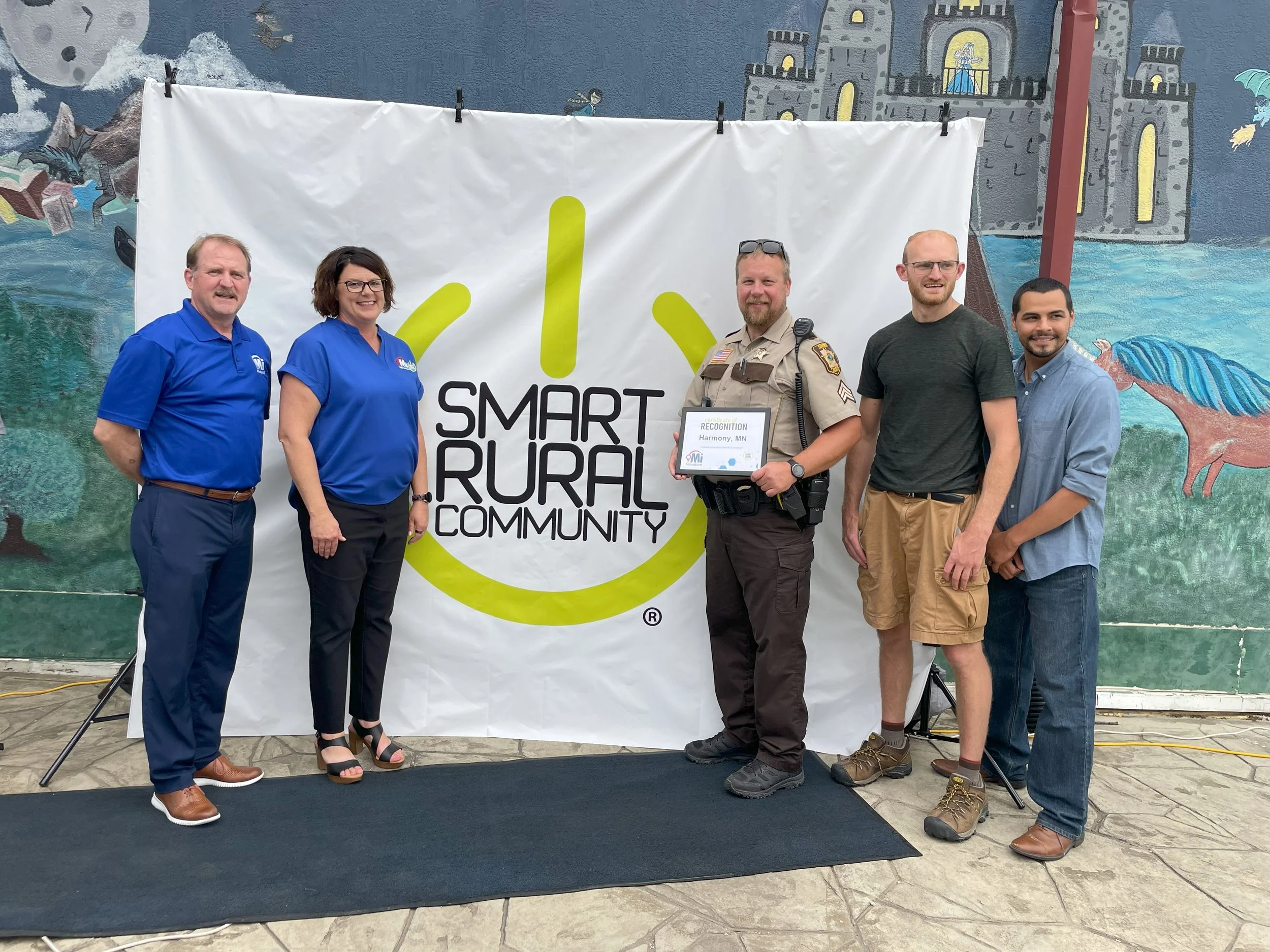 Four people standing in front of a white background with the words "Smart Rural Communities" written on it. The group includes three women and one man, all smiling and dressed in business casual attire.