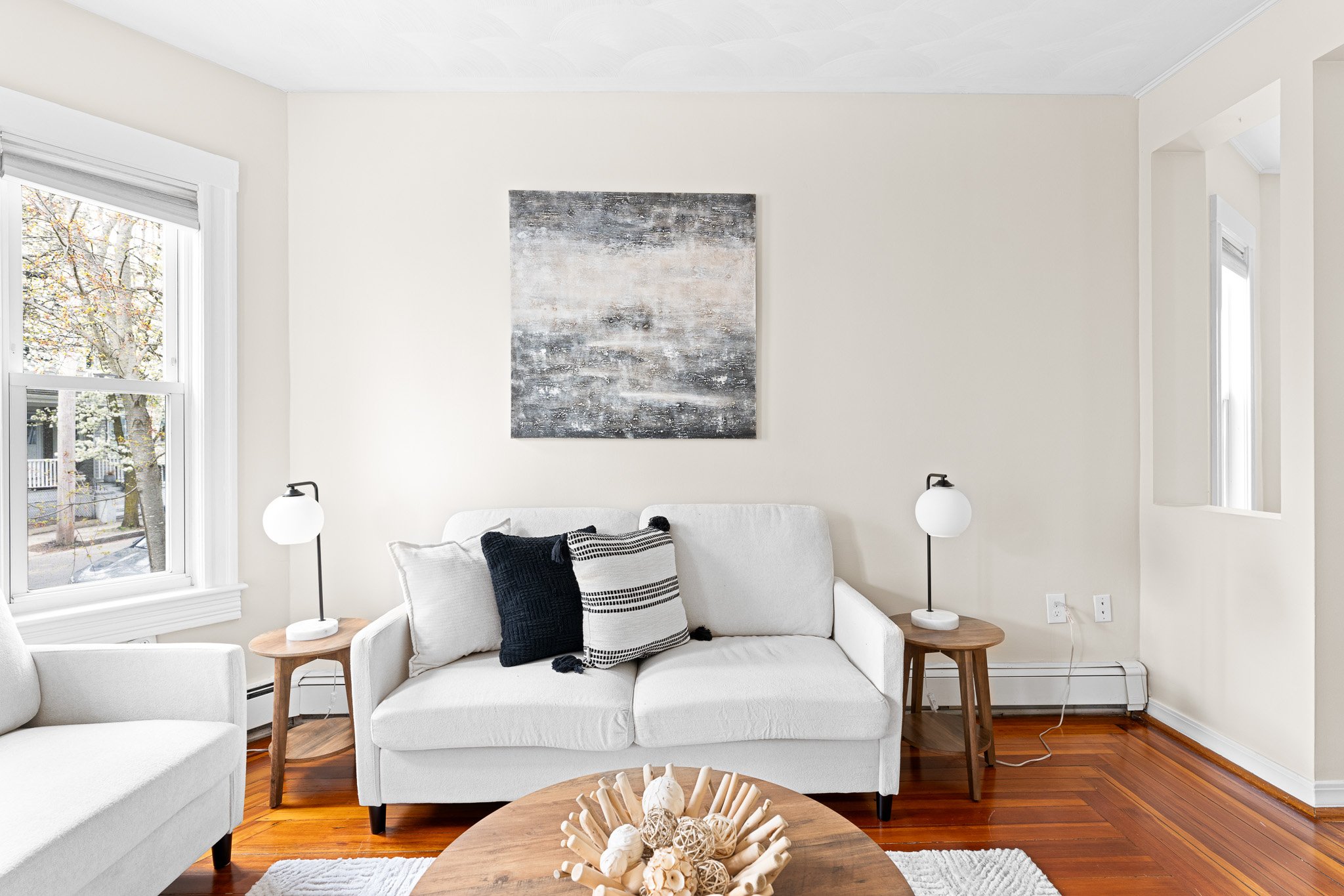 Living room with white sofa, black and white pillows, two side tables with white lamps, wooden coffee table decorated with natural beige decor, wood flooring, and large windows with blinds.