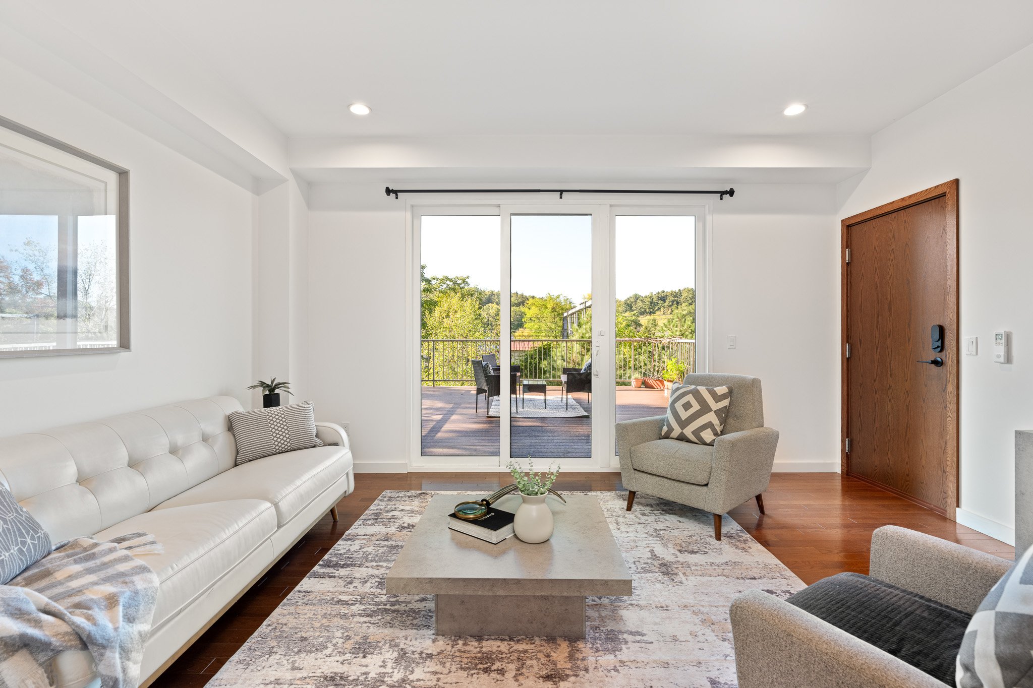 Living room with white sofa, gray armchair, coffee table with flowers, large sliding glass doors leading to outdoor patio, and views of trees.