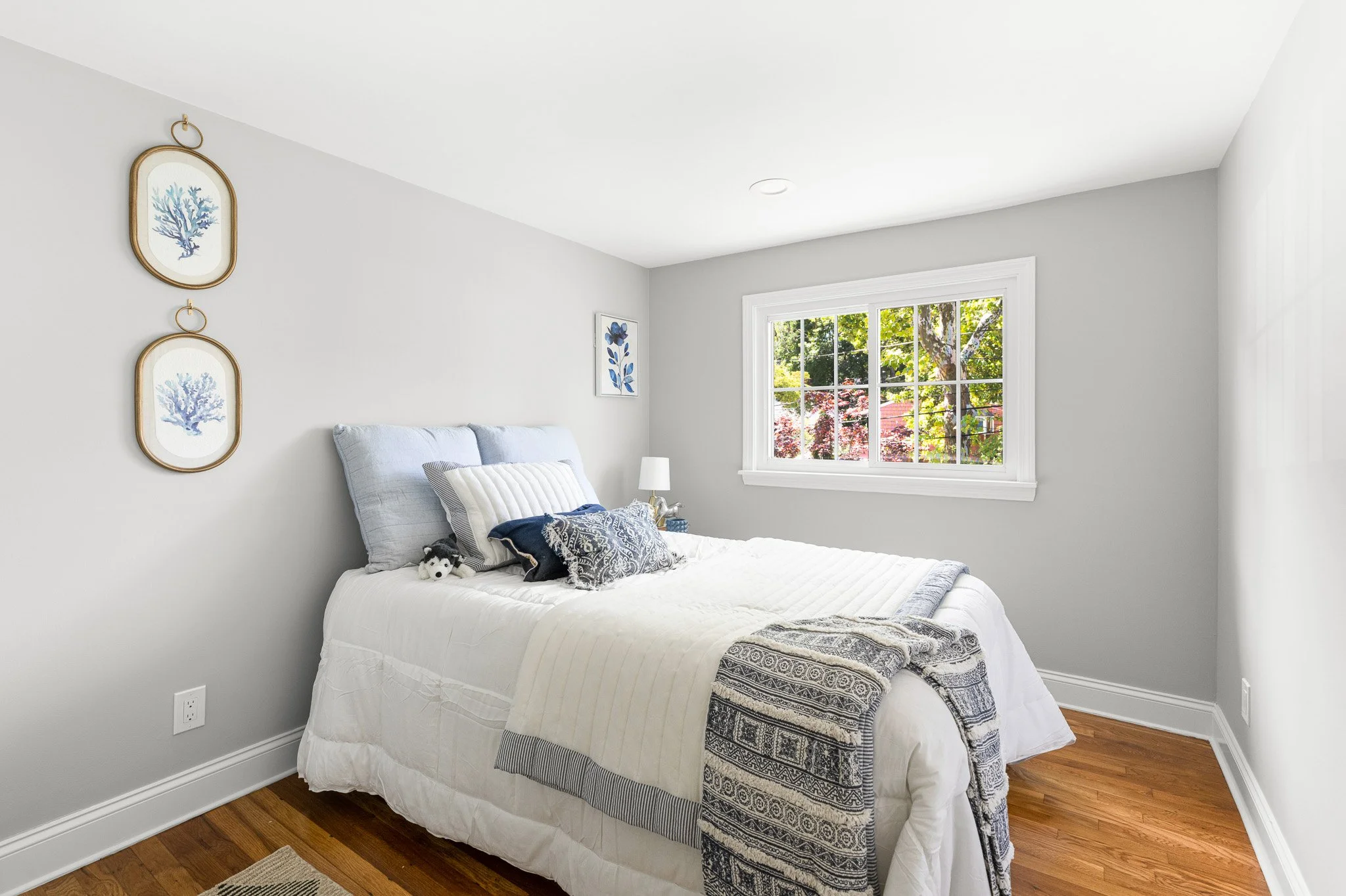 A bright bedroom with white walls, a large window with greenery outside, a bed with a white quilt, pillows, and a small stuffed animal on it, and wall art featuring blue coral images.