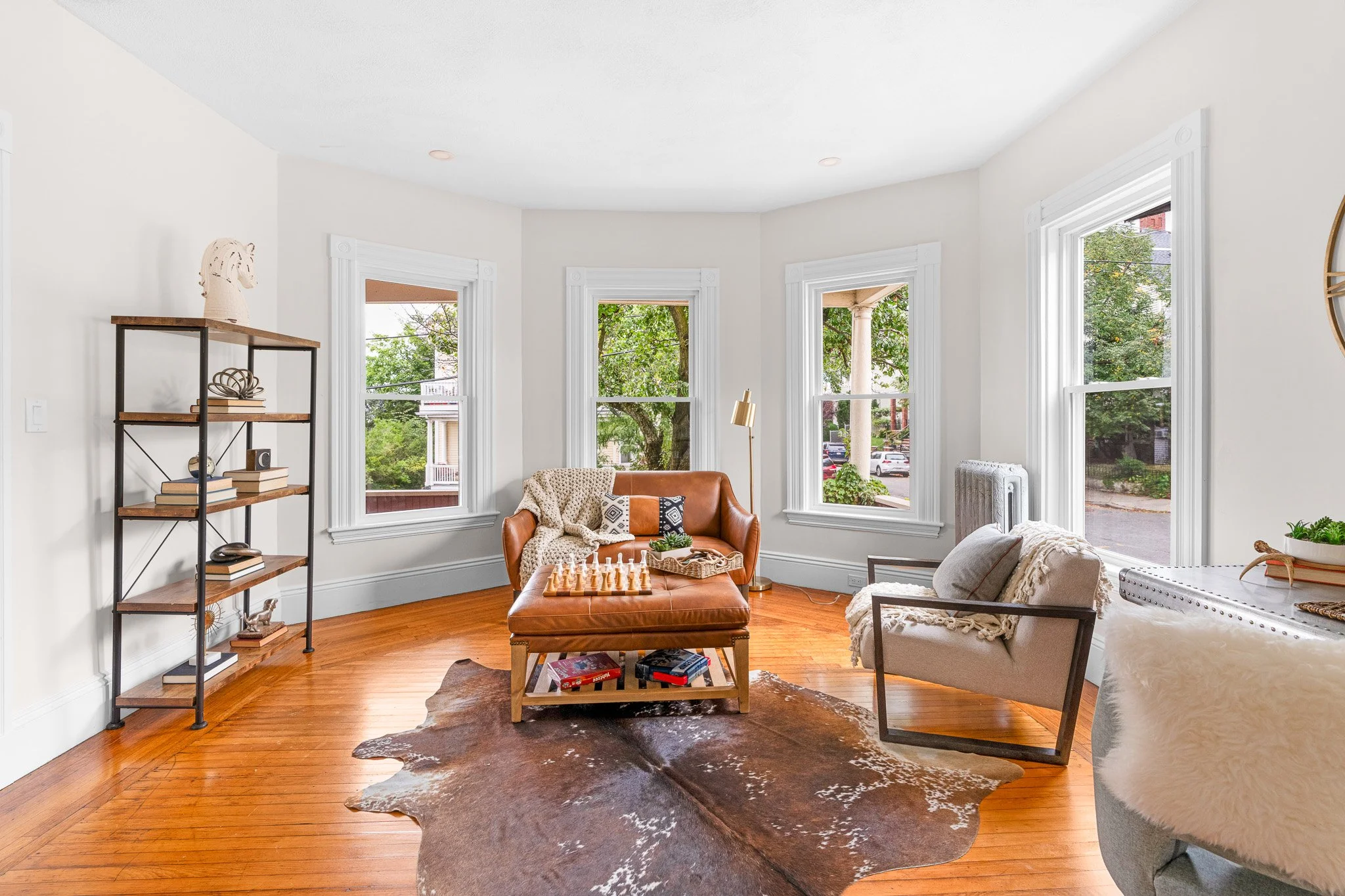 Bright living room with multiple large windows, a leather ottoman with a chess set, a cream-colored sofa, a white armchair, a black metal shelving unit with decorative items, a cowhide rug, and a wooden floor.