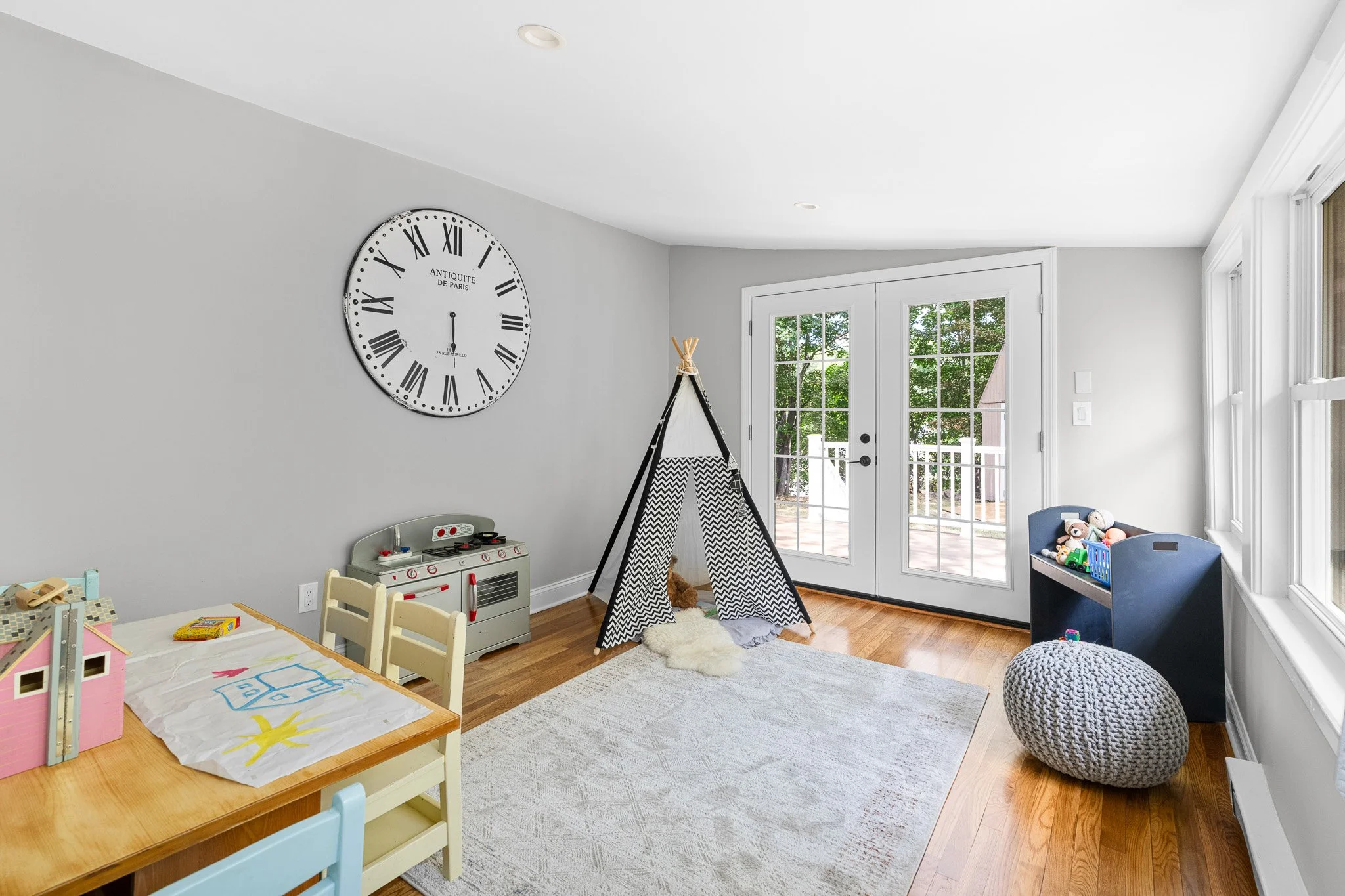 A cozy children's playroom with light gray walls, a large wall clock, a teepee tent, a shelf of toys, a wooden table with chairs, and a view of the backyard through glass doors.