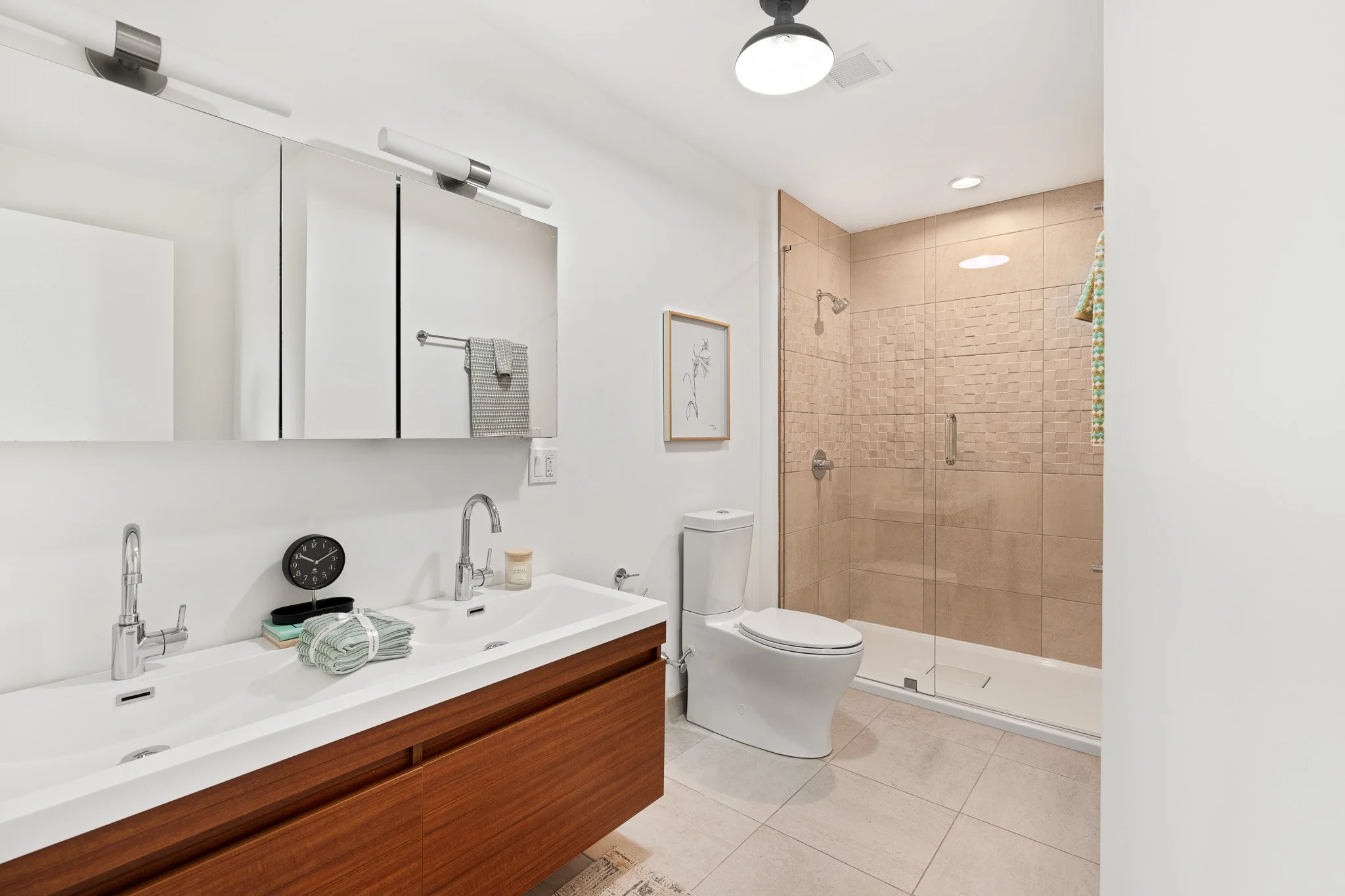 Modern bathroom with white walls, a double vanity with wood cabinetry, a towel rack with gray towels, a round wall clock, a framed botanical picture, a toilet, and a glass-enclosed shower with beige tiles.