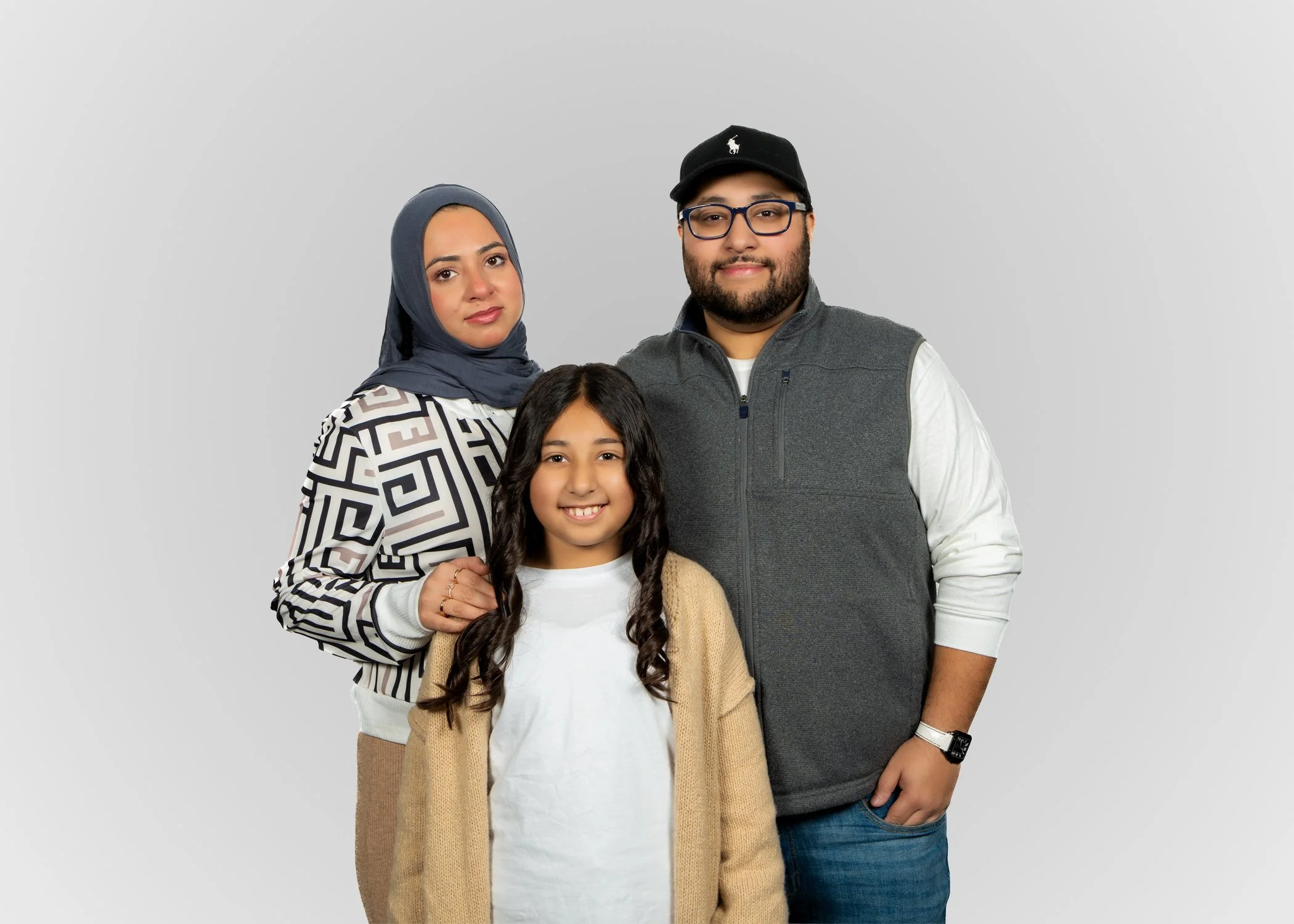 A family of four posing together against a plain gray background. The family includes a woman wearing a headscarf, a man with glasses and a cap, a young girl with long dark hair, and a woman with black hair, all smiling and standing close.