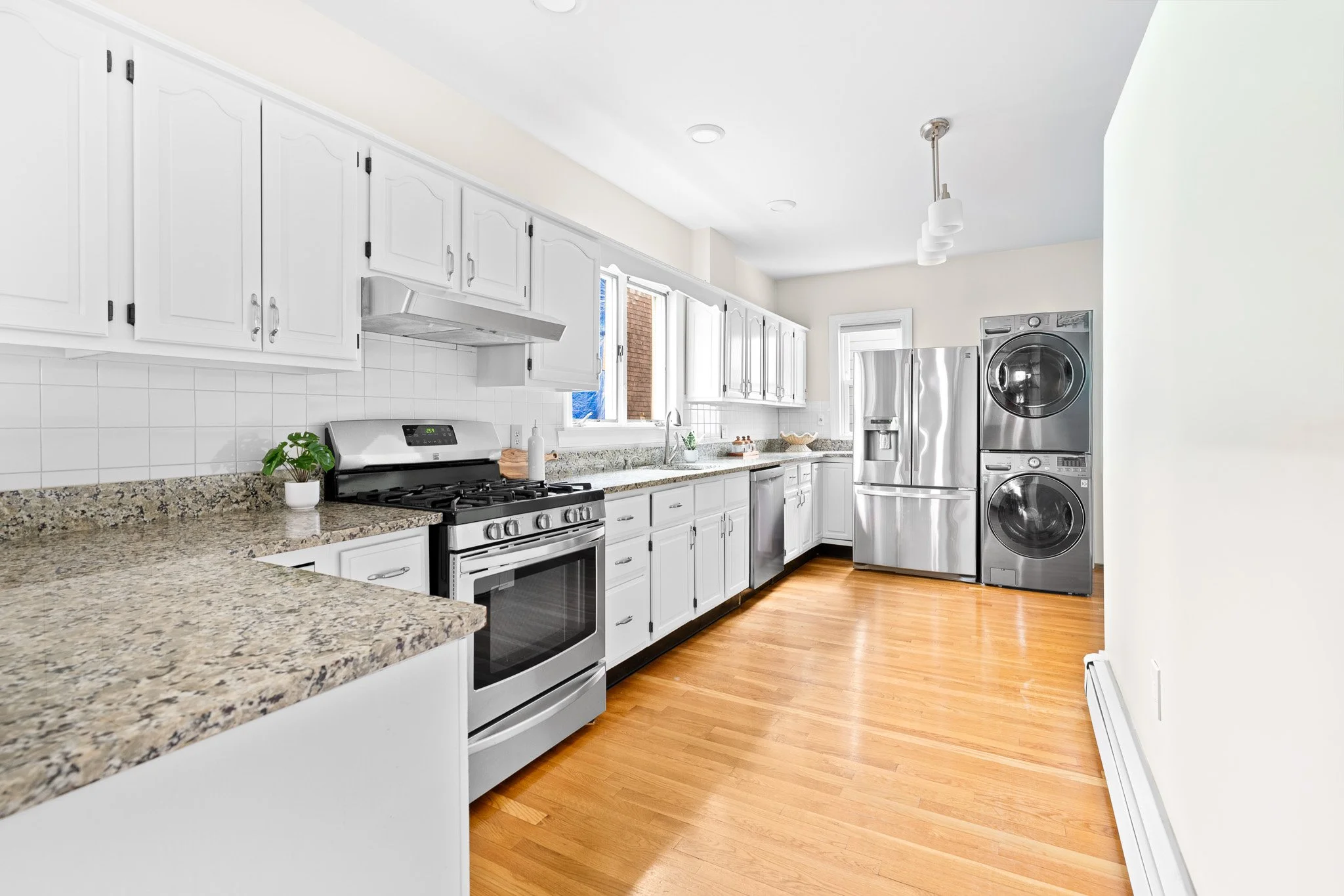 A kitchen with white cabinets, a granite countertop, stainless steel appliances including a stove, refrigerator, and stacked washer and dryer, and a hardwood floor.