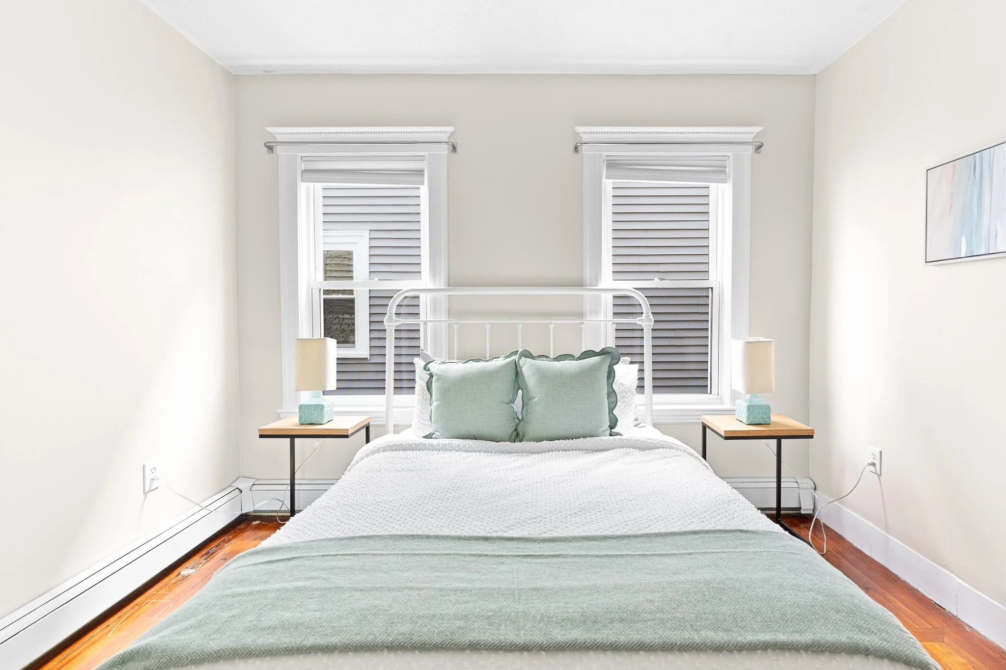 A minimalist bedroom with a white bed and pale green pillows, two wooden nightstands with lamps, and two windows with shutters.