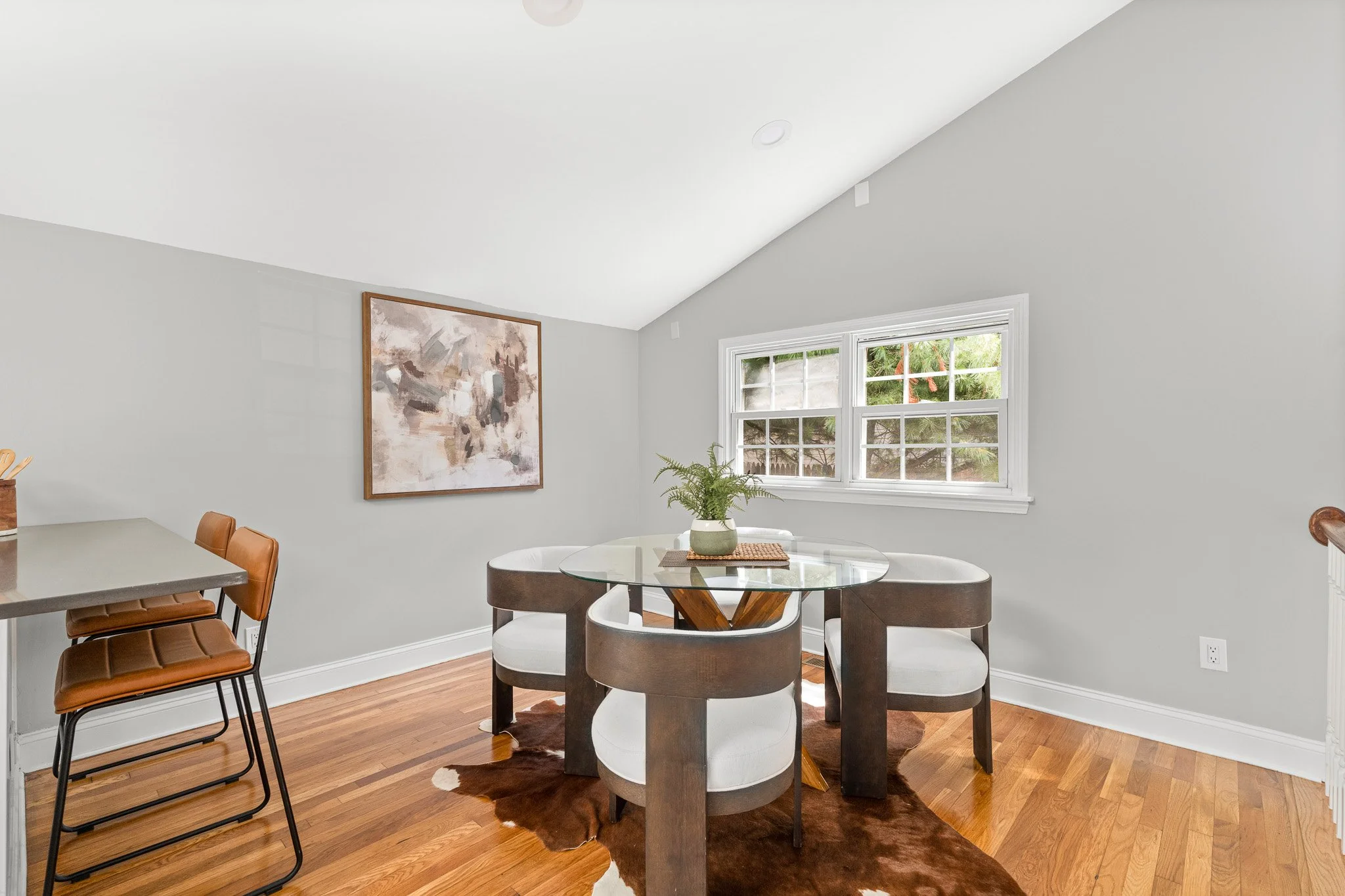 A dining room with a round glass table, four white and dark brown chairs, a cowhide rug, a window with greenery outside, a modern abstract painting on the wall, a small plant on the table, and wooden flooring.