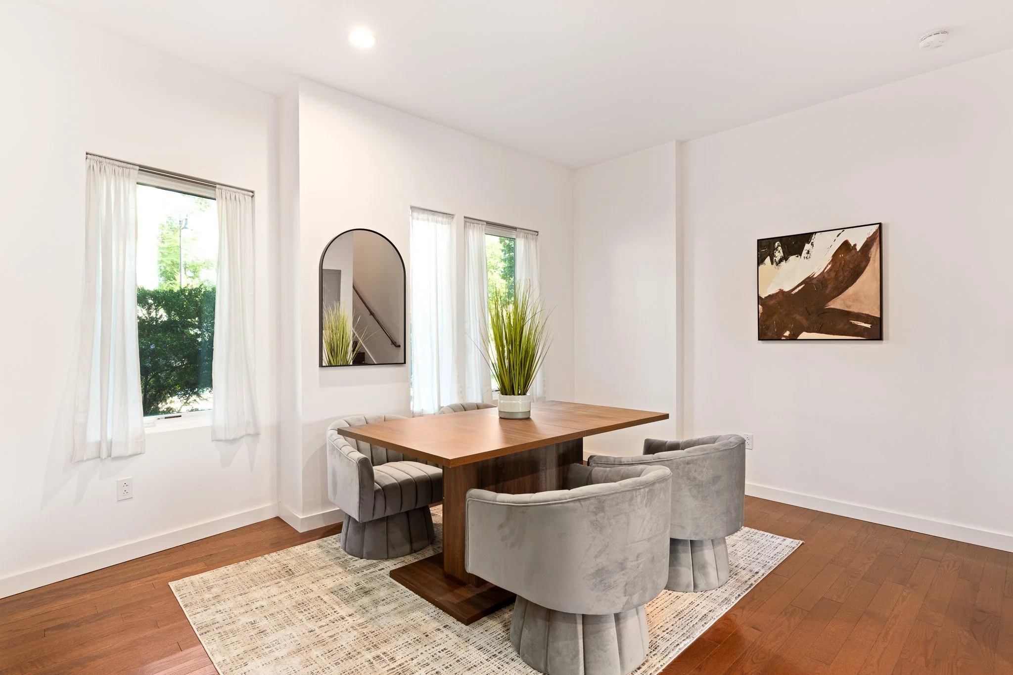 A modern dining room with white walls, three windows with sheer white curtains, a wooden dining table, and four upholstered chairs. There is a potted plant on the table and a framed abstract painting on the wall. An arched mirror reflects part of a staircase.