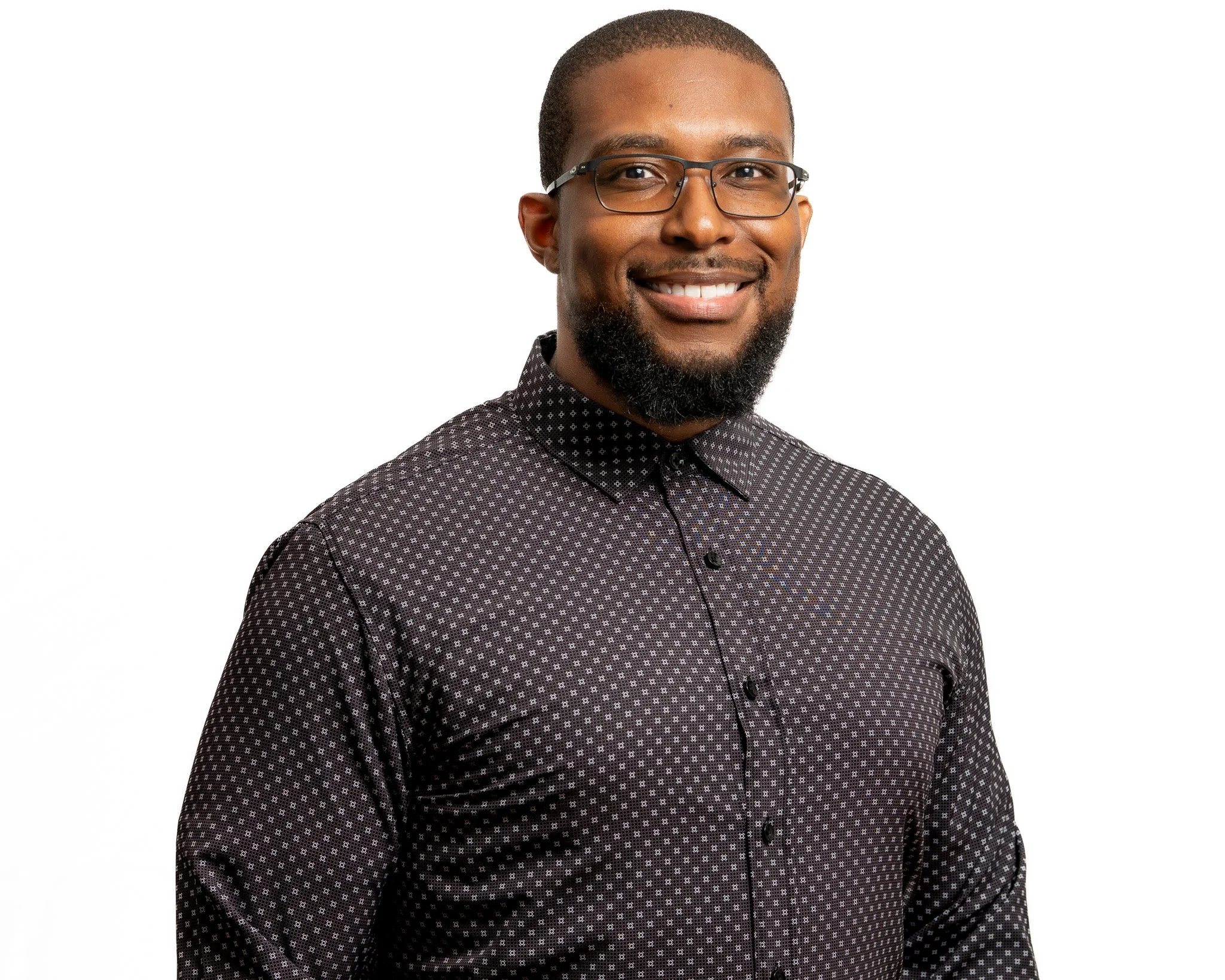 A smiling African American man with glasses, a beard, and a dark patterned shirt, standing against a white background.