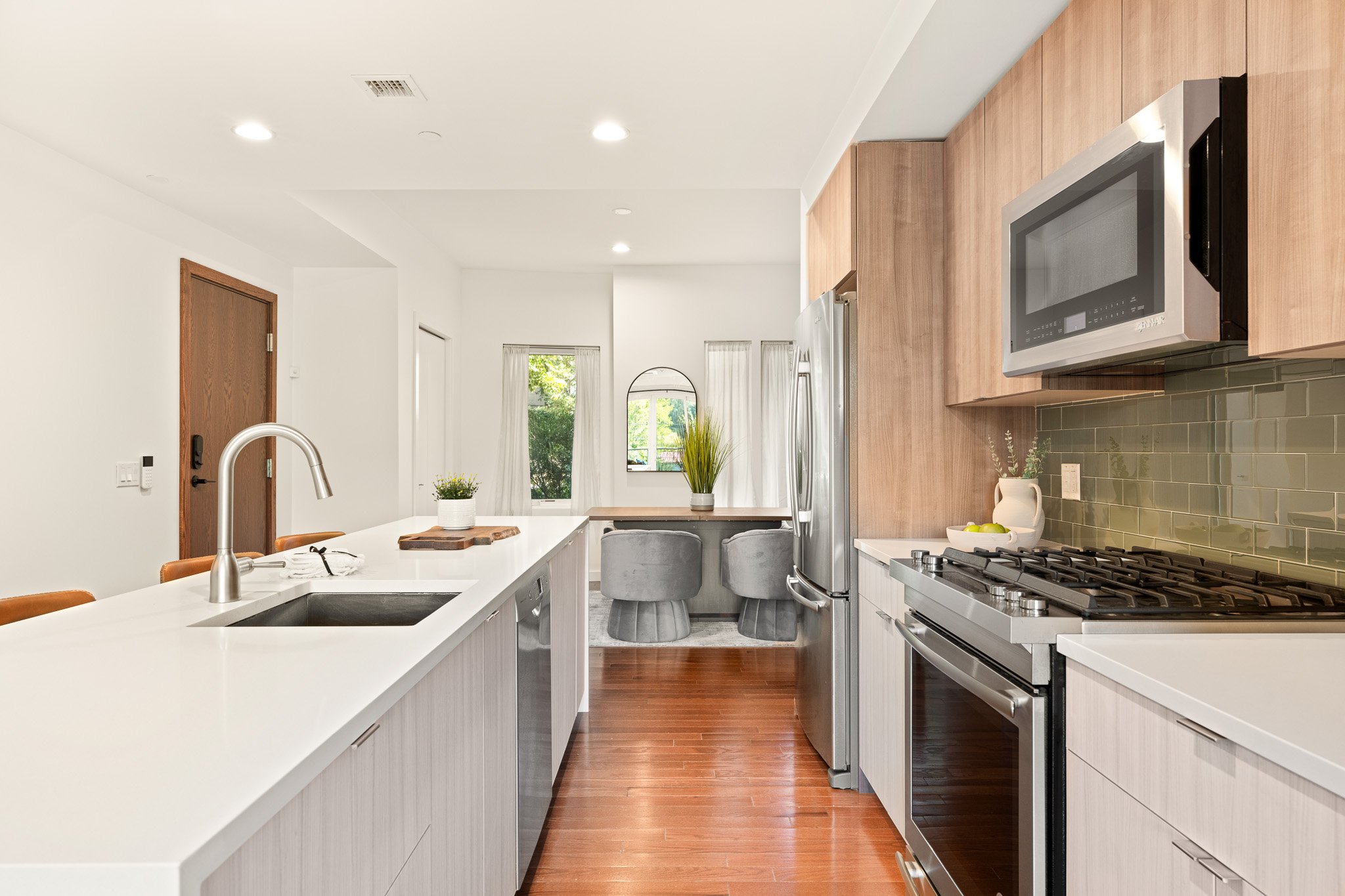 A modern kitchen with white countertops, wooden cabinetry, stainless steel appliances, and a dining area with gray chairs, large windows with white curtains, and greenery outside.