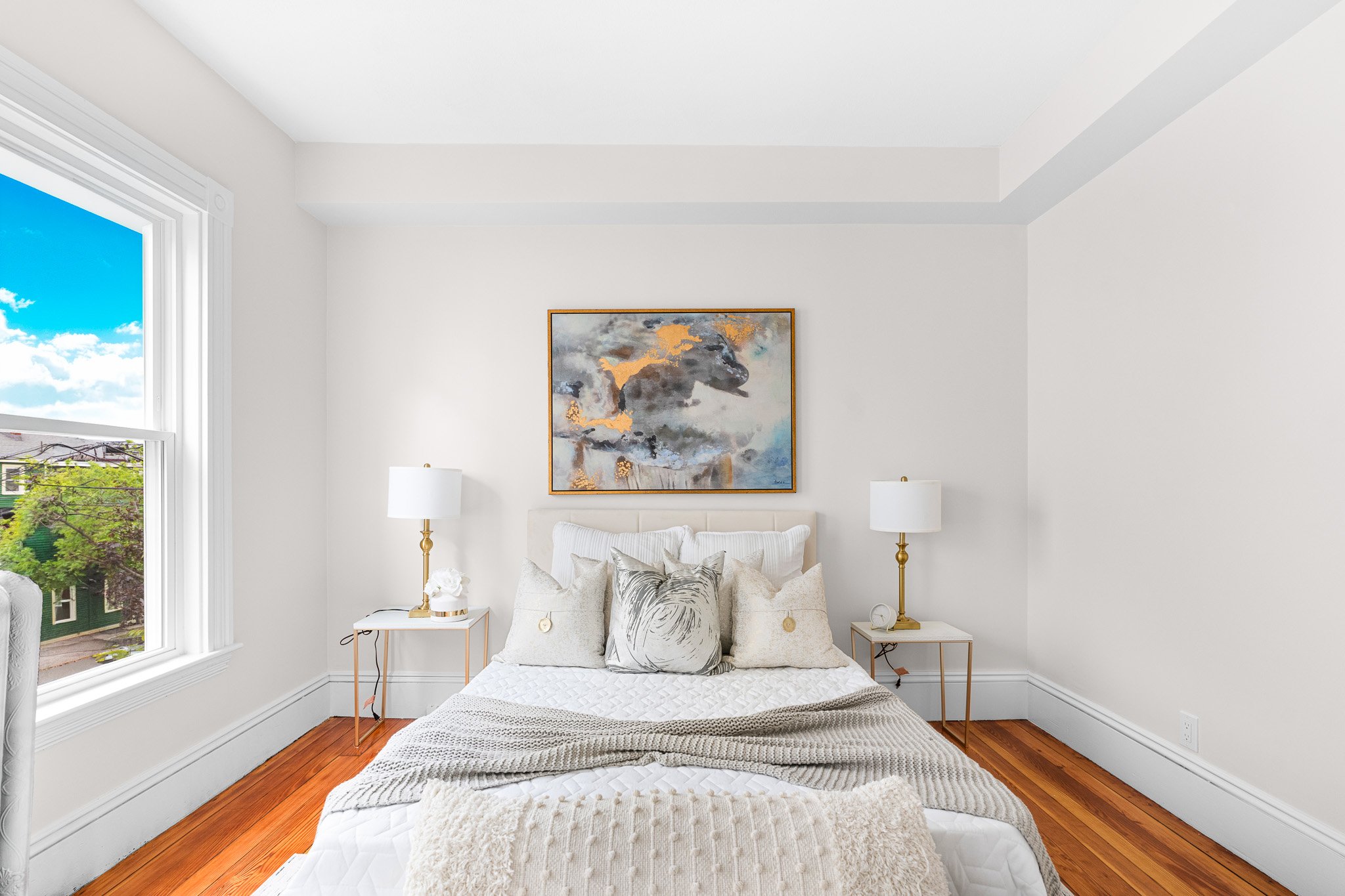 A bedroom with white walls and a window showing a cloudy sky and trees outside. There is a white headboard with decorative pillows, two white nightstands with gold lamps, and an abstract painting of a horse hanging above the bed.