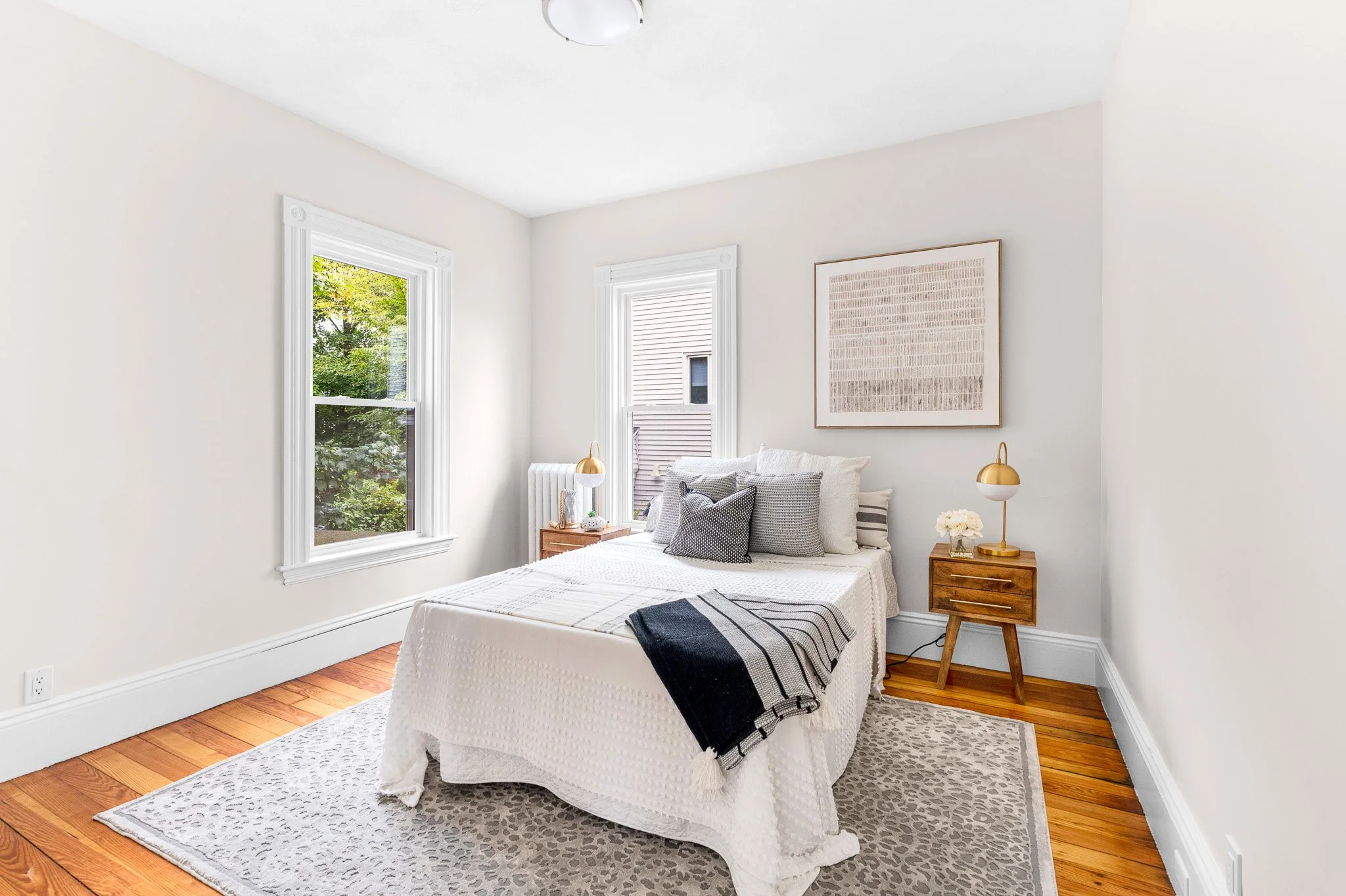 A bright bedroom with two windows, a white bed with textured bedding, black and white pillows, a wooden nightstand with a gold lamp and flowers, a framed artwork on the wall, a beige patterned rug, and hardwood floors.