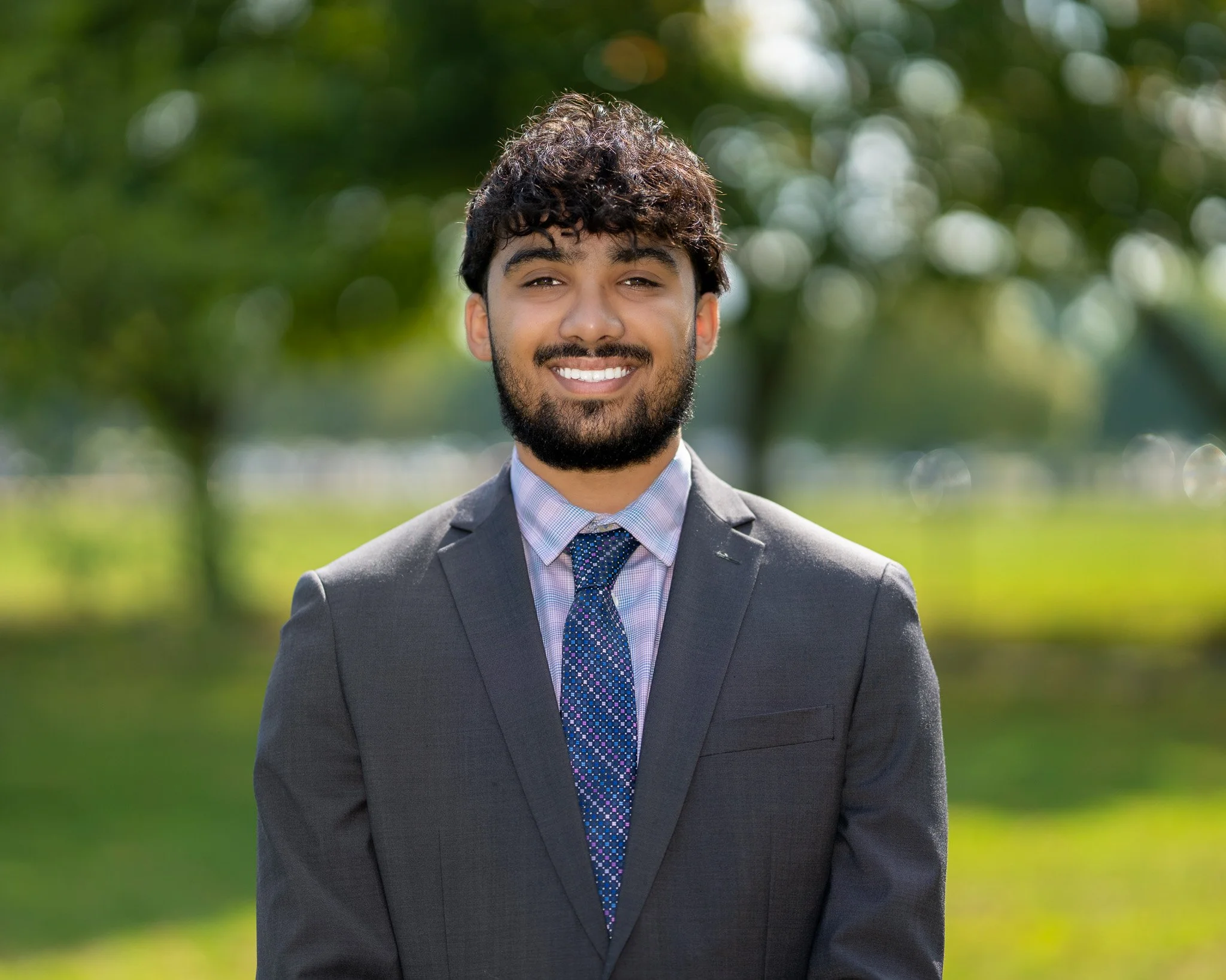 A man with dark curly hair, a beard, and a suit smiling outdoors with a blurred green and white bokeh background.