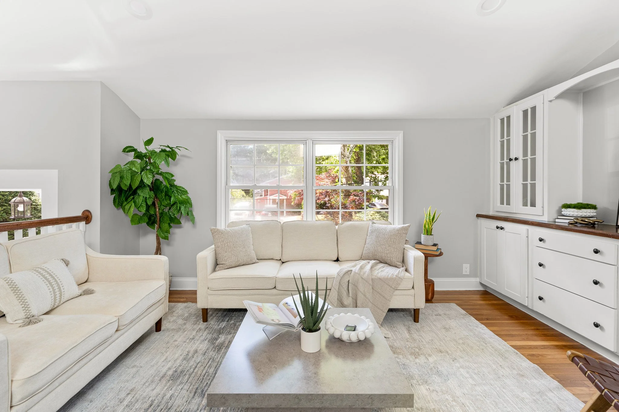 Living room with white sofas, large window showing green trees outside, potted plants, and a coffee table with an open book and decorative items.