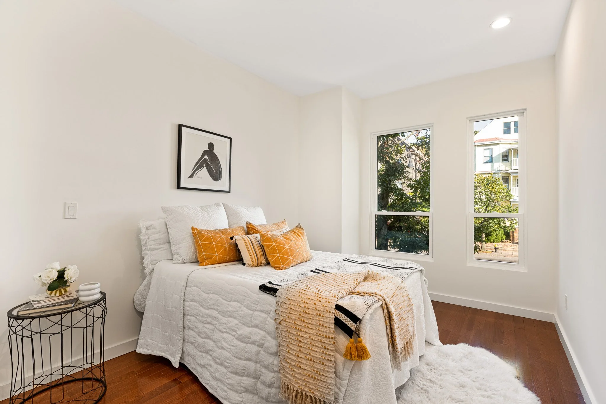 A cozy bedroom with a bed covered in white bedding and orange pillows. Next to the bed, there is a small black metal side table with decorative plants and books. The wall behind the bed has one framed black and white minimalist art print. Two large windows let in natural light and show a view of trees and neighboring houses. The room features white walls, wooden floors, and a fluffy white rug.