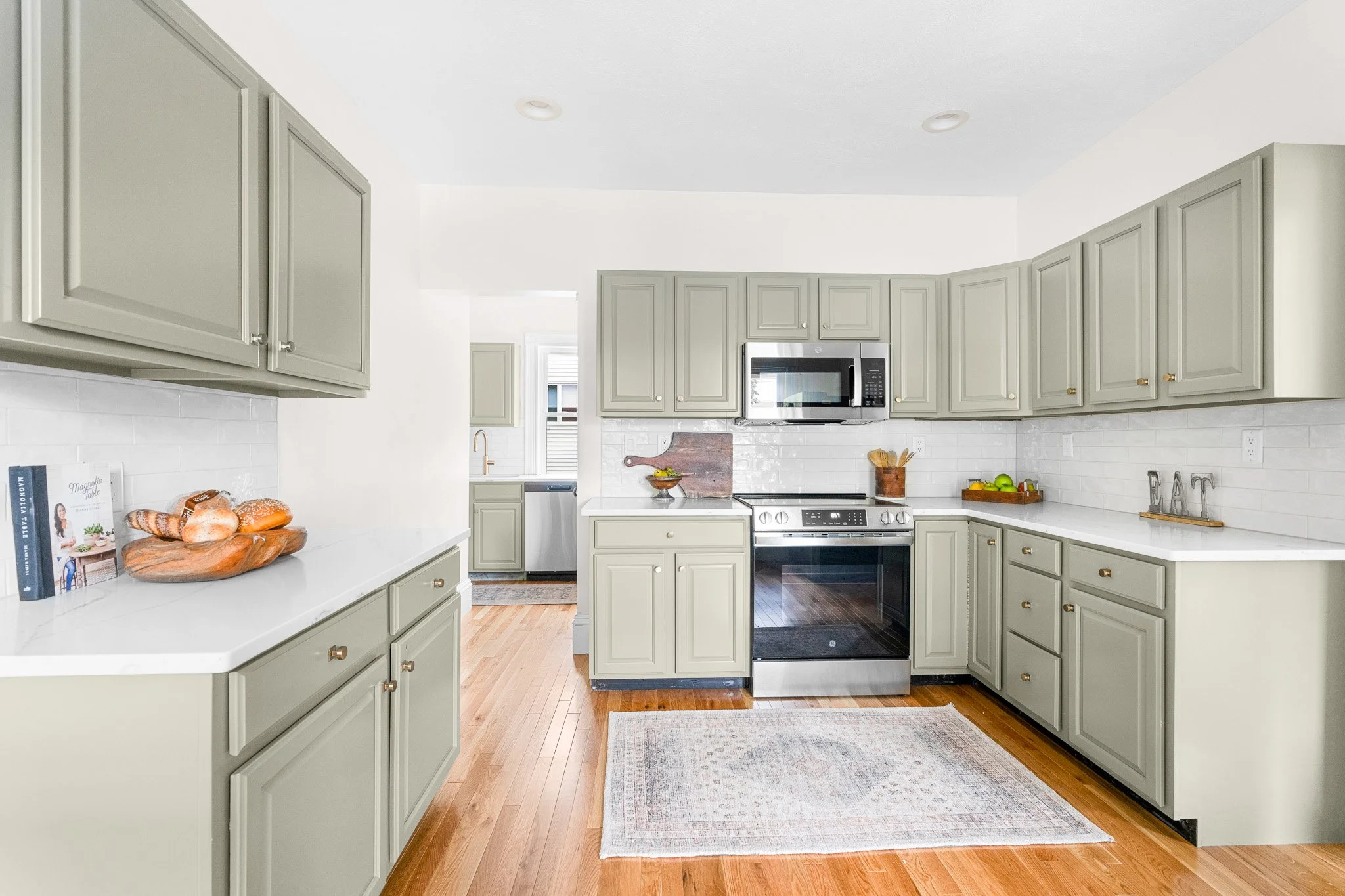 A kitchen with light green cabinets, white countertops, a white backsplash, stainless steel appliances, and wooden floors.