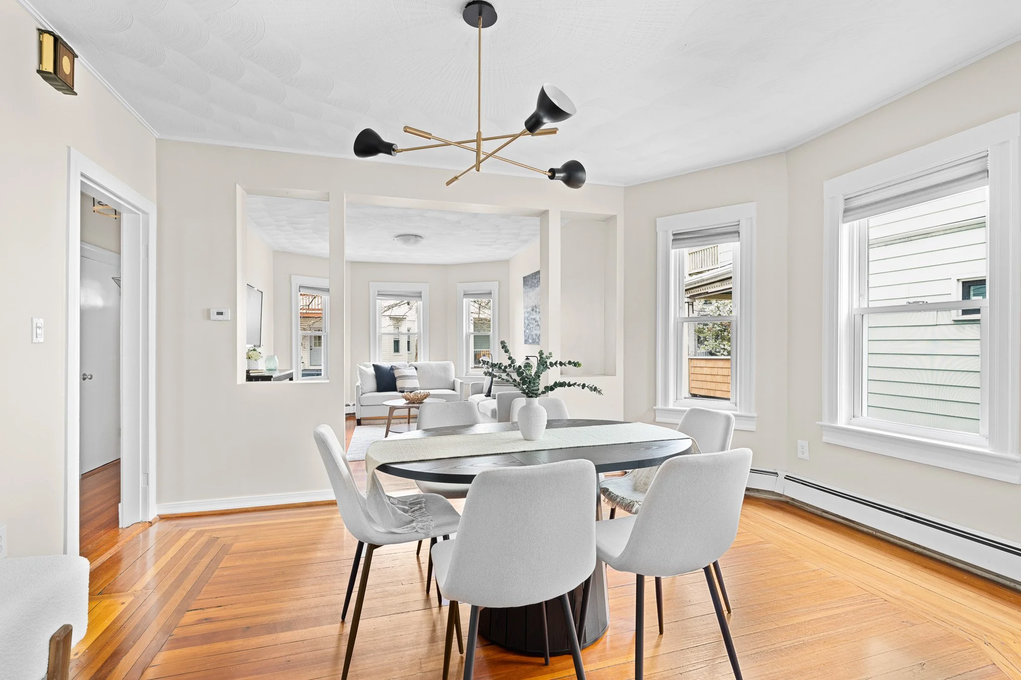 A bright dining room with white walls and large windows. There is a black round dining table with white chairs around it. The room is decorated with a white vase and green plant on the table, and a modern black and gold chandelier hanging from the ceiling. Visible in the background is a living area with white sofa and artwork.