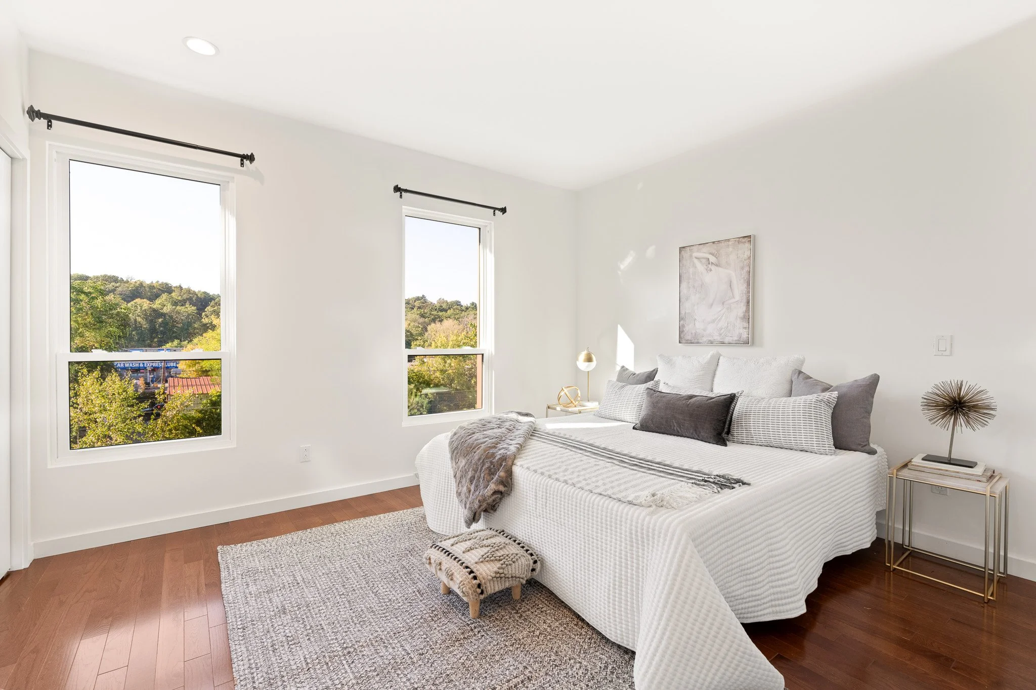 Modern bedroom with a white bed, gray and white pillows, a throw blanket, a bedside table with a golden lamp, artwork on the wall, windows showing a view of trees, and hardwood floors.
