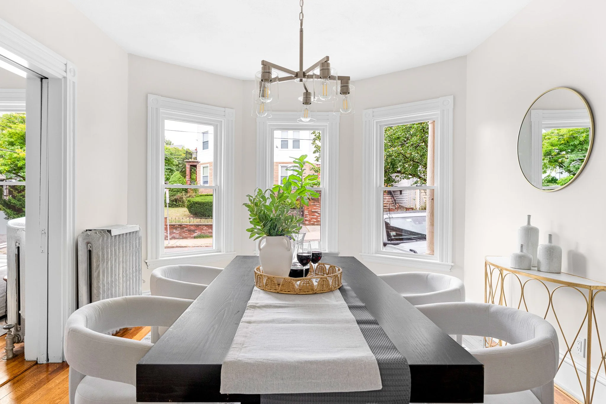 A dining room with a dark wooden table, white upholstered chairs, a centerpiece with a white vase and wine glasses, large windows with greenery outside, a gold-colored side table with white vases, and a modern chandelier hanging from the ceiling.