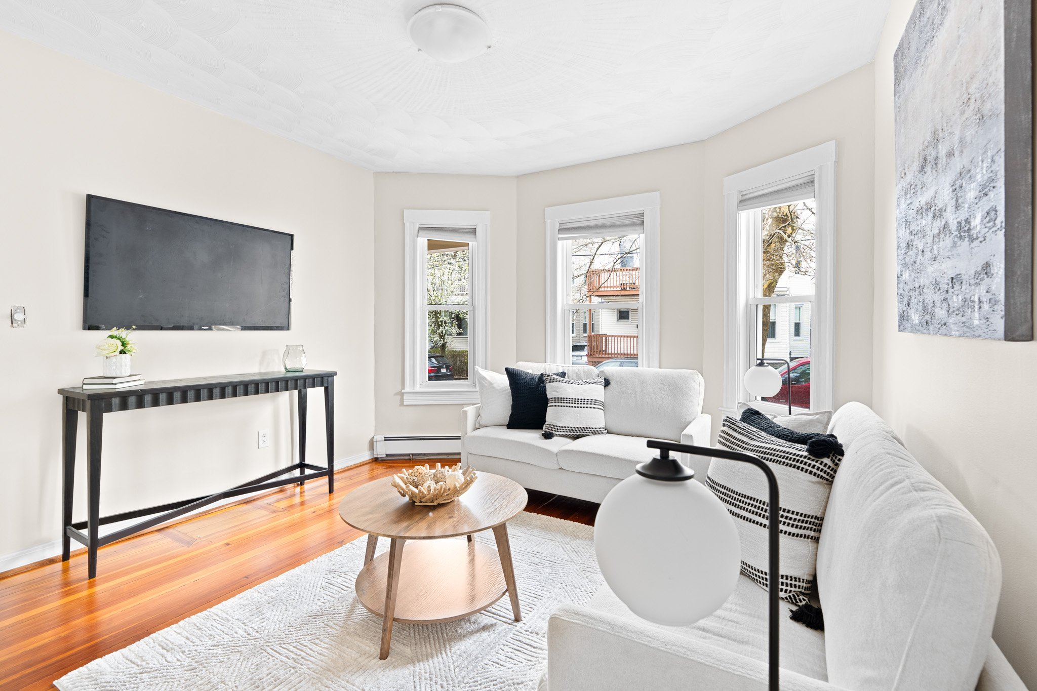 Living room with white walls, three large windows, a mounted flat-screen TV, a white sofa with black and white pillows, a round wooden coffee table with seashell decor, a white round table lamp, a wooden console table, and a textured wall art piece.