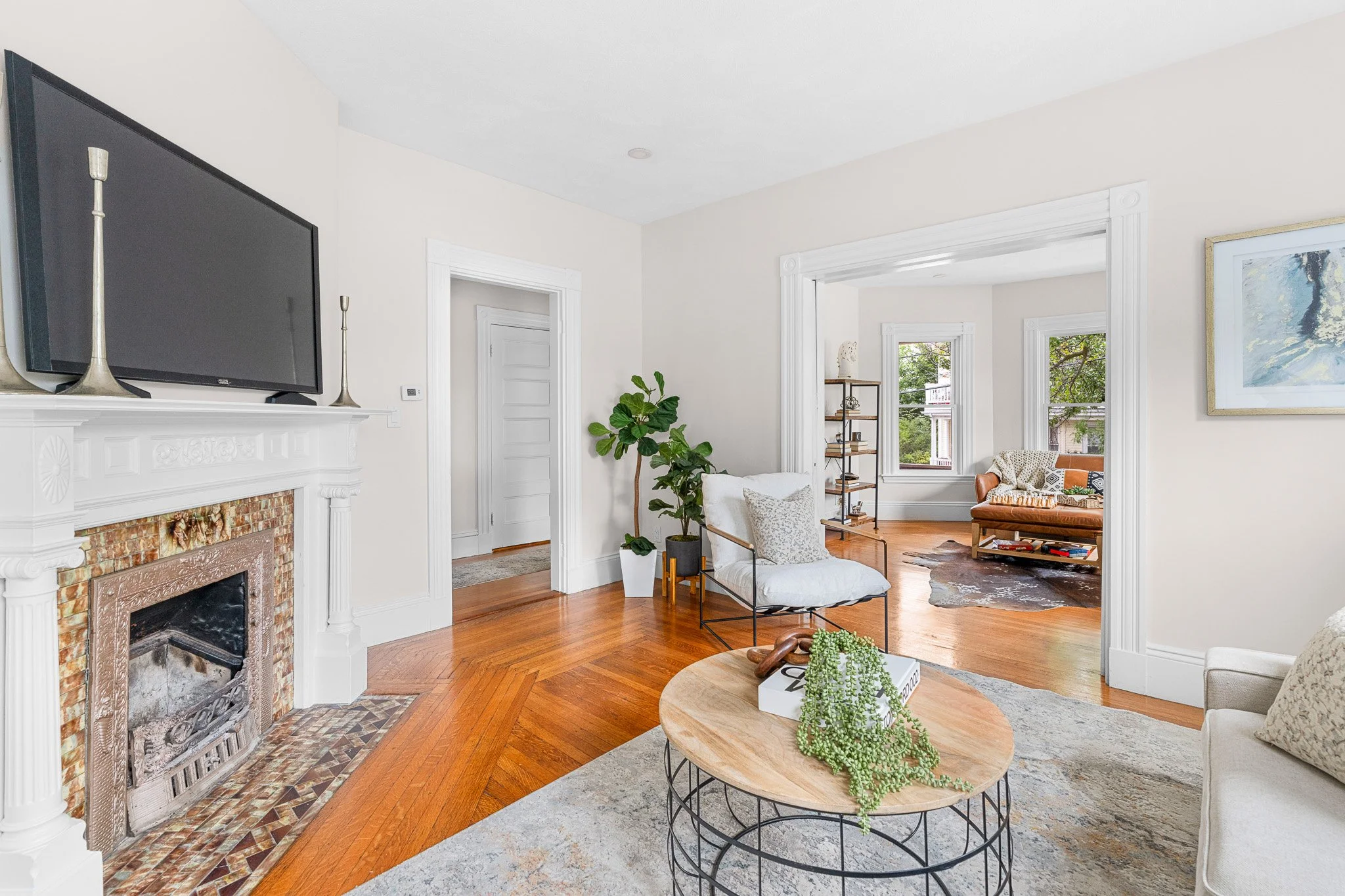 A bright living room with a fireplace, a wall-mounted flat-screen TV, a cozy armchair, a coffee table with a plant, and large windows showing green trees outside.