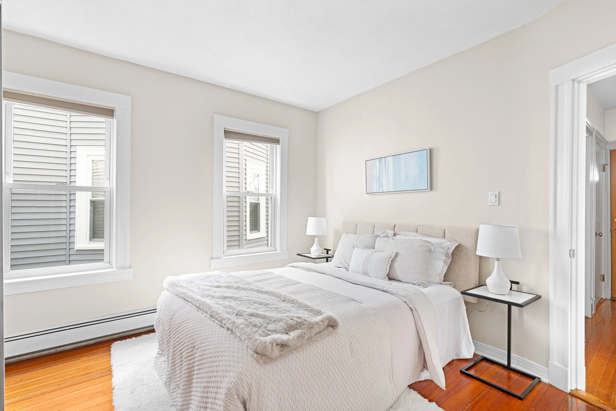 Bright bedroom with white walls, two windows with blinds, a white bed with textured bedding, and nightstands with white lamps.