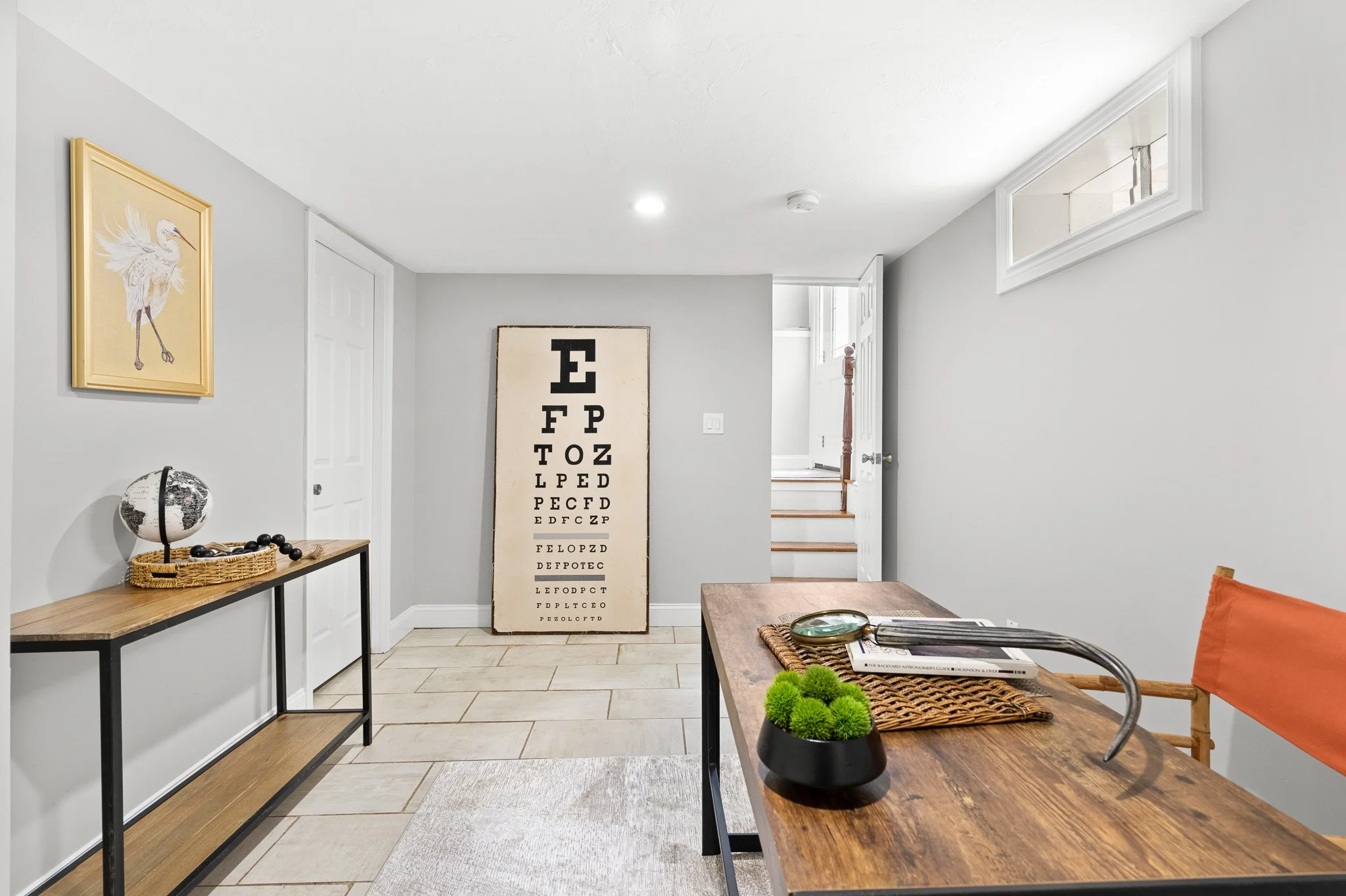 Living room with gray walls, wood and metal side table with globe, eye chart leaning against wall, and desk with orange pillow, silver globe, black beads, and potted green plants.