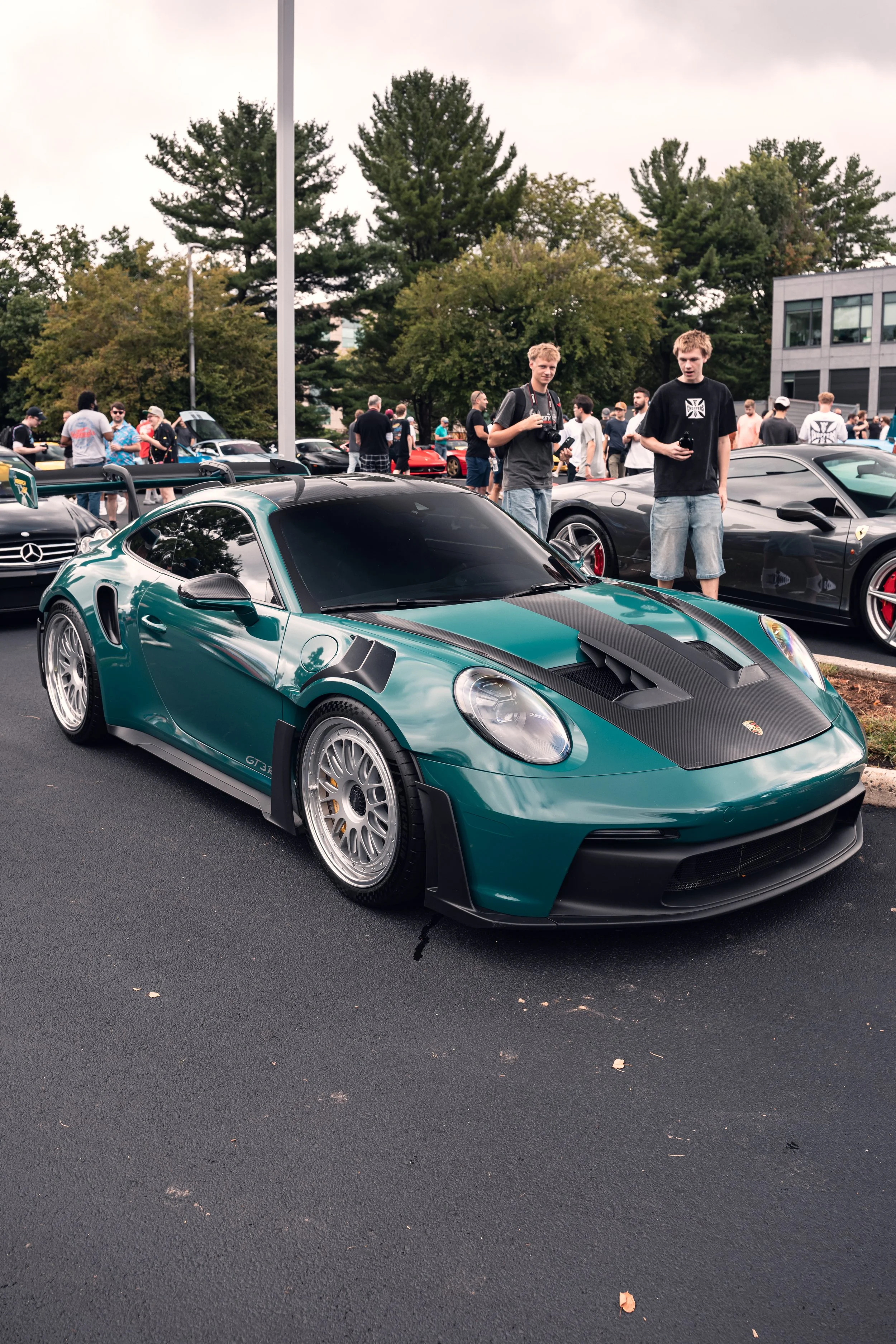 A green sports car with black accents and large wheels is parked at a car show along with other vehicles. Two young men are standing nearby, one holding a camera and the other holding a phone, looking at the camera. Several people are in the backgrou