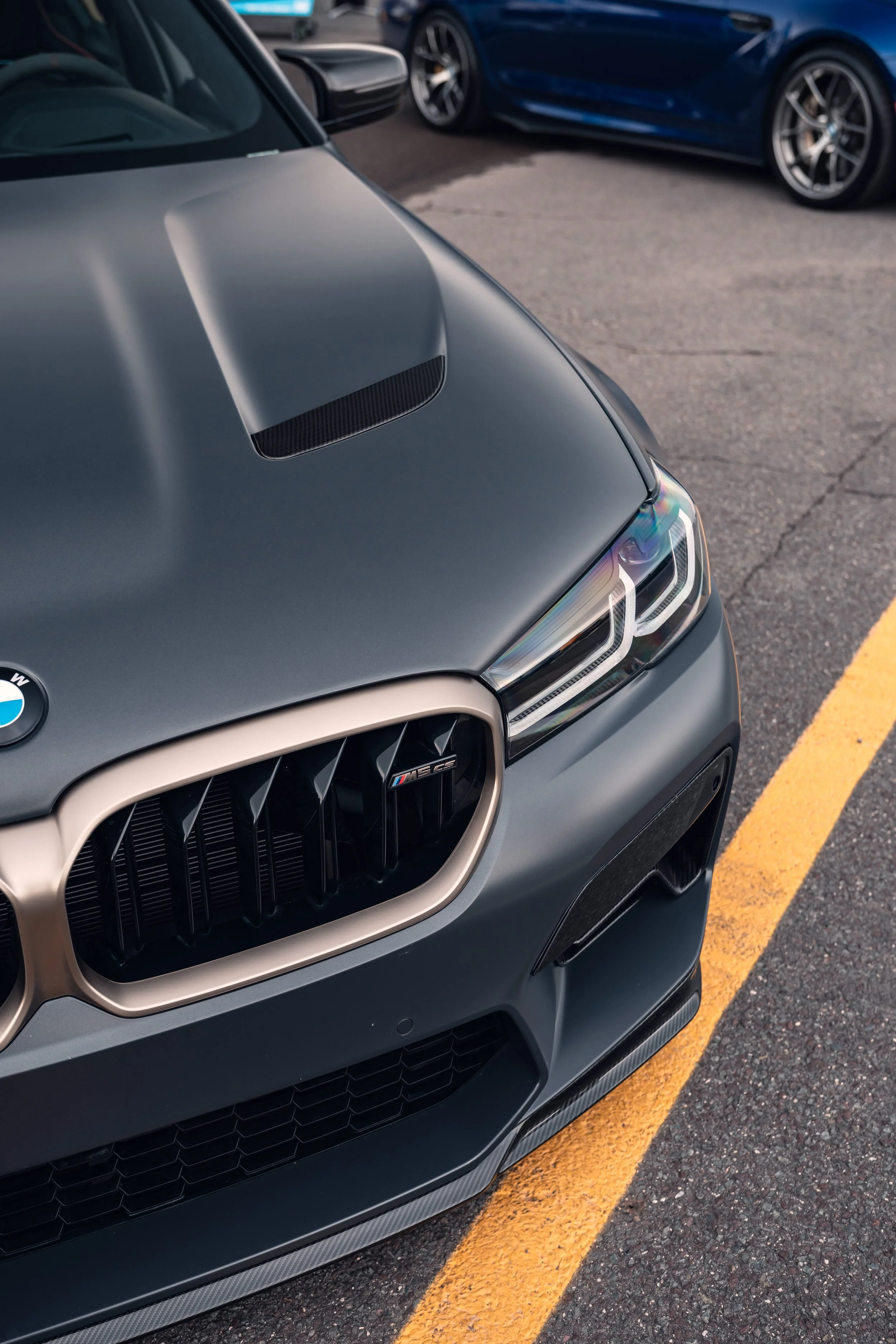 Close-up of the front of a gray BMW M8 GTS sports car parked on a street, with a blue sports car in the background.