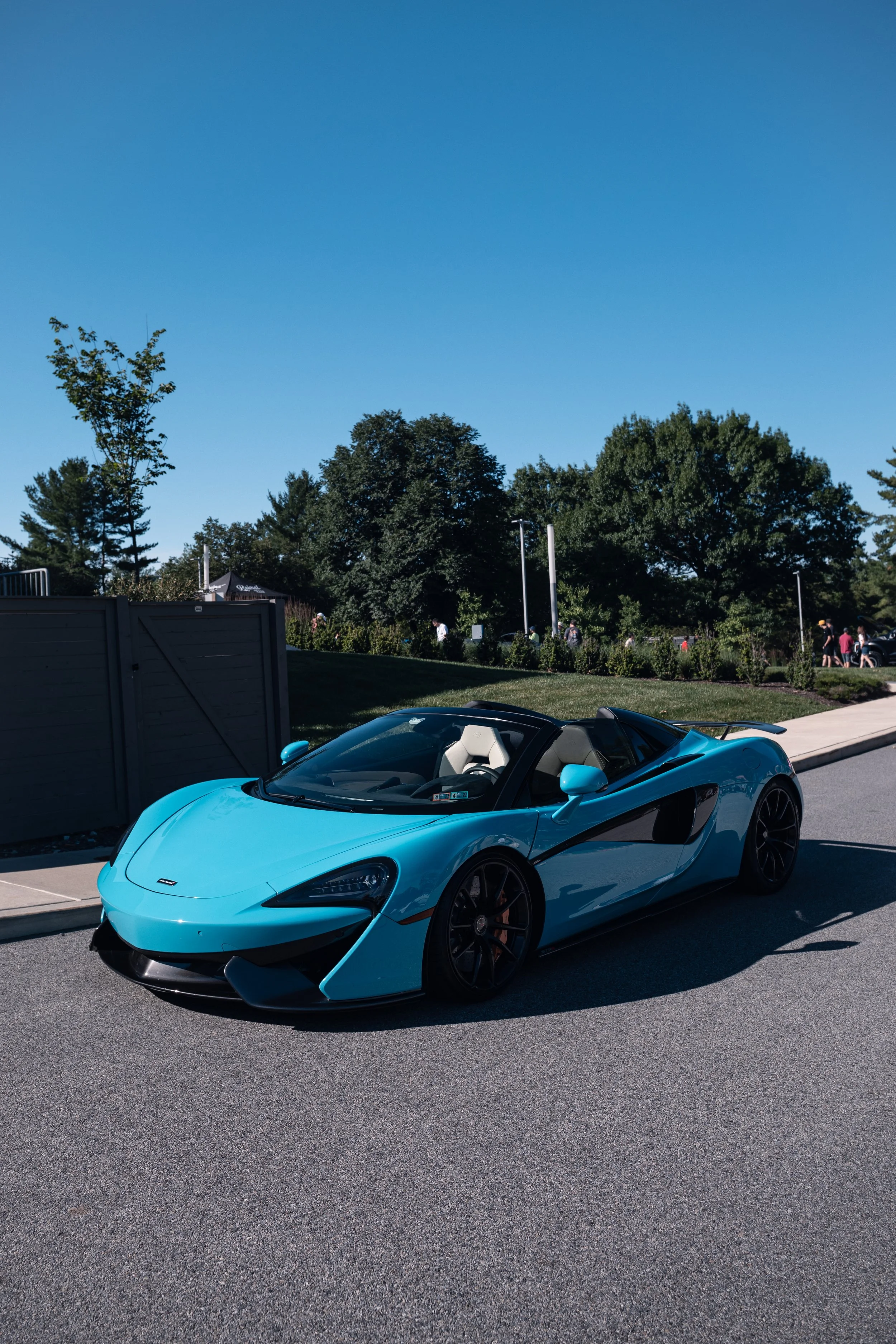 A blue sports convertible car parked on a sunny day with a background of green trees, people walking, and a clear blue sky.