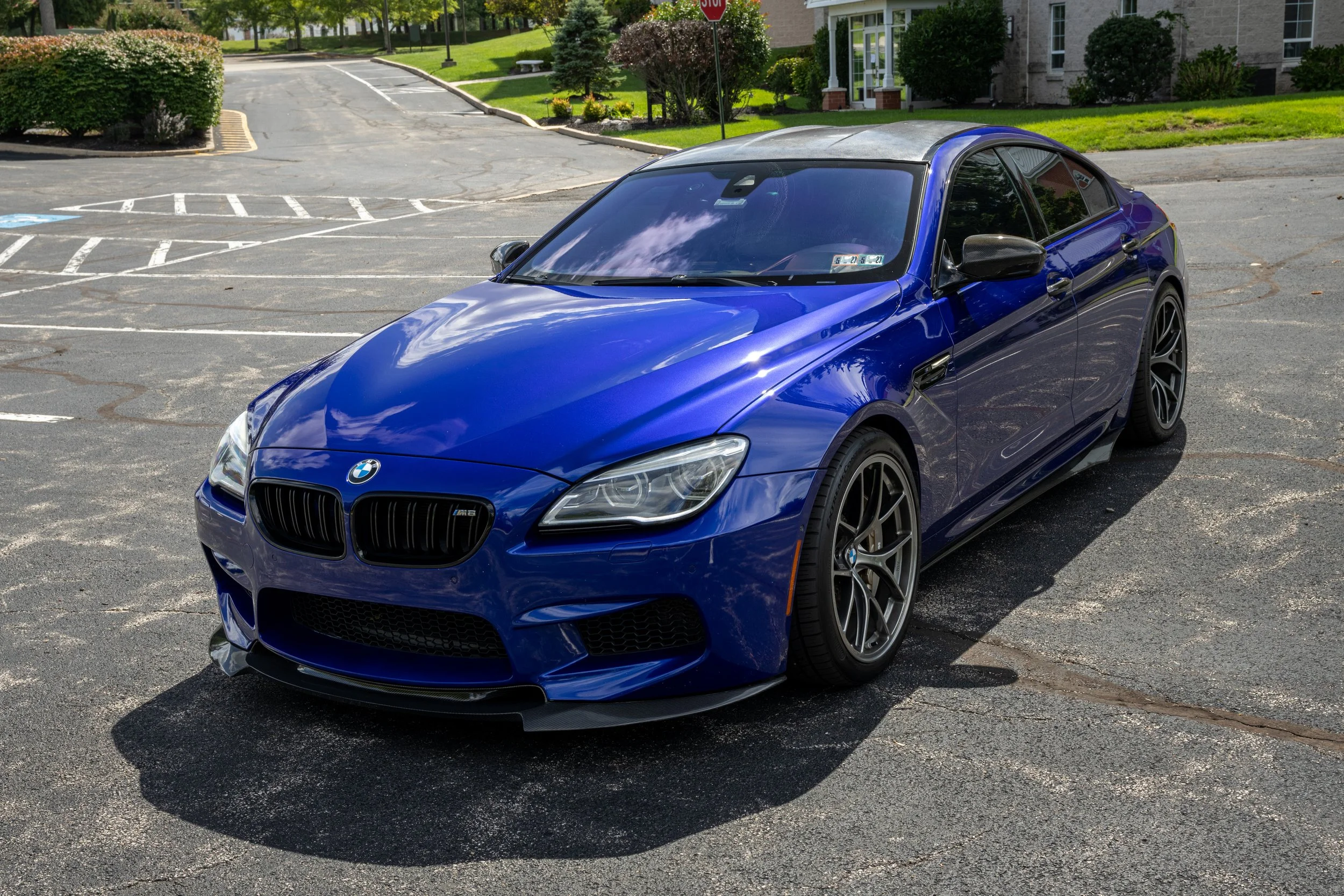 A blue BMW M8 sports car parked in an empty parking lot with white line markings and a handicap parking space. In the background, there are green bushes, trees, and a residential building with windows and a porch.